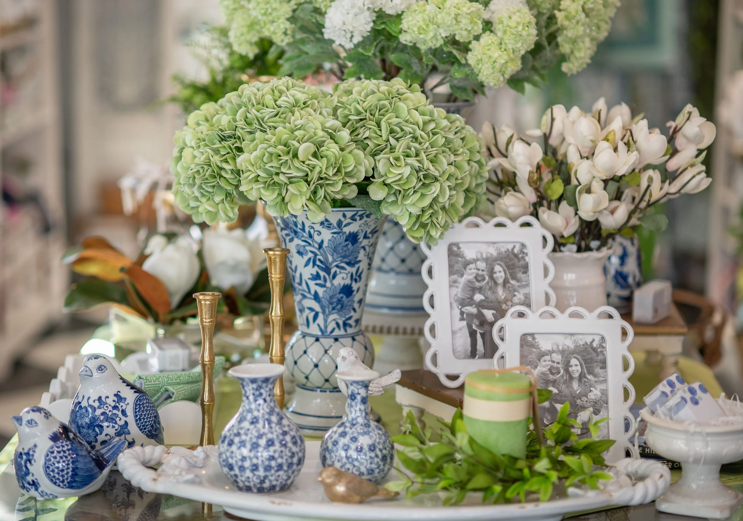 Arrangement of green hydrangeas in blue and white porcelain vases, along with porcelain figurines and picture frames on a table.