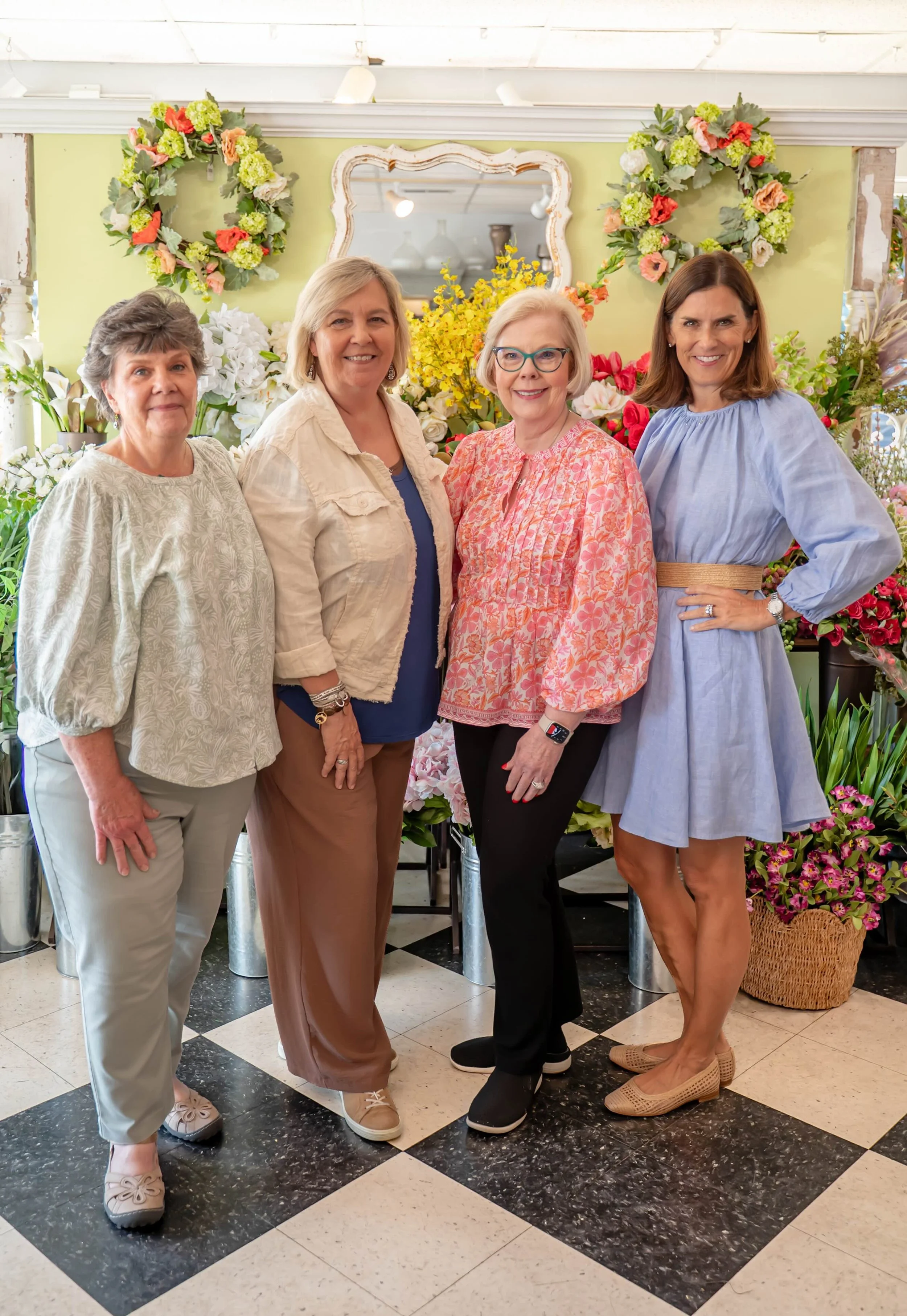 Four women standing together in a floral shop, surrounded by colorful flowers and wreaths, smiling at the camera.