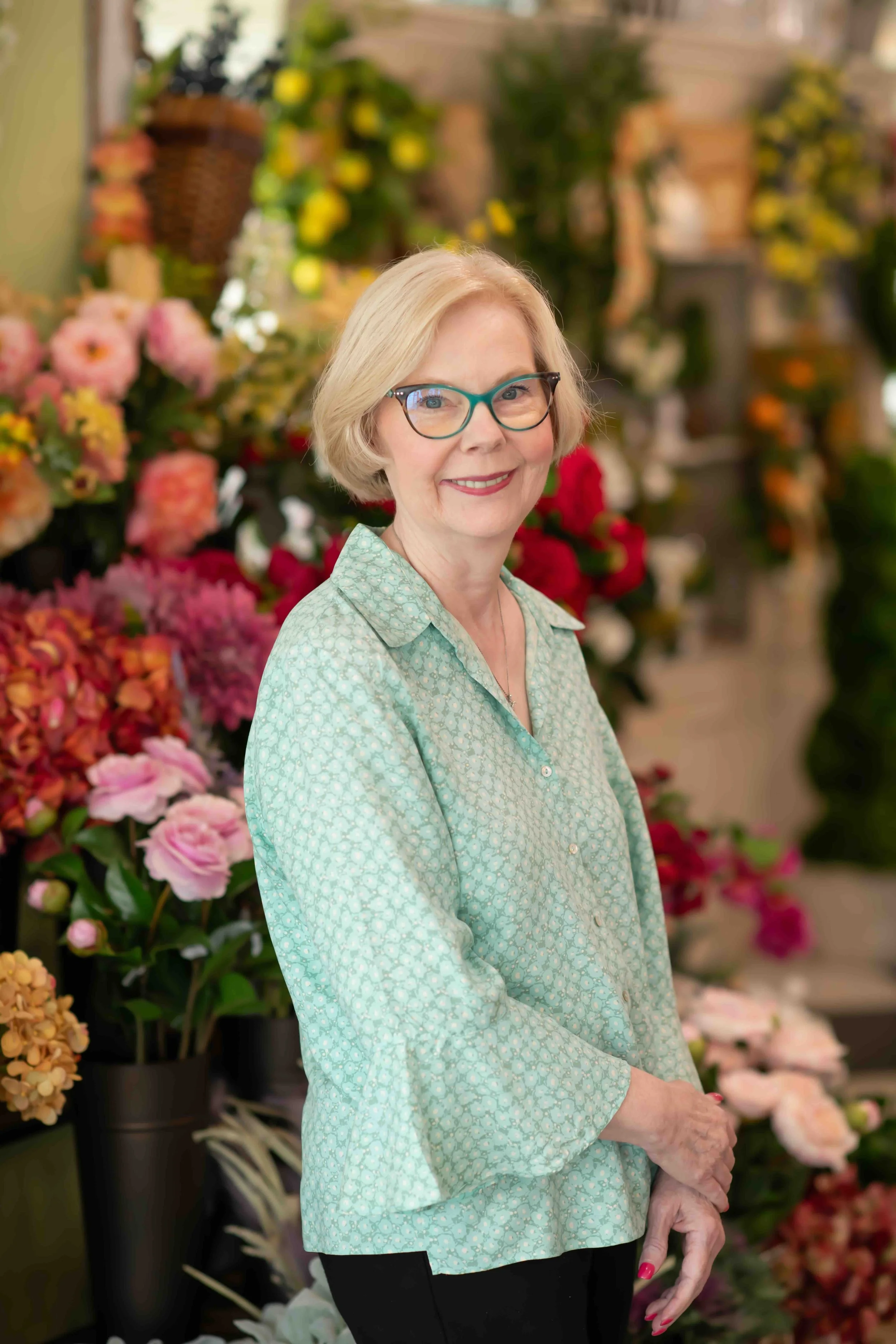 A smiling woman with blonde hair and glasses standing in front of colorful flowers in a flower shop.