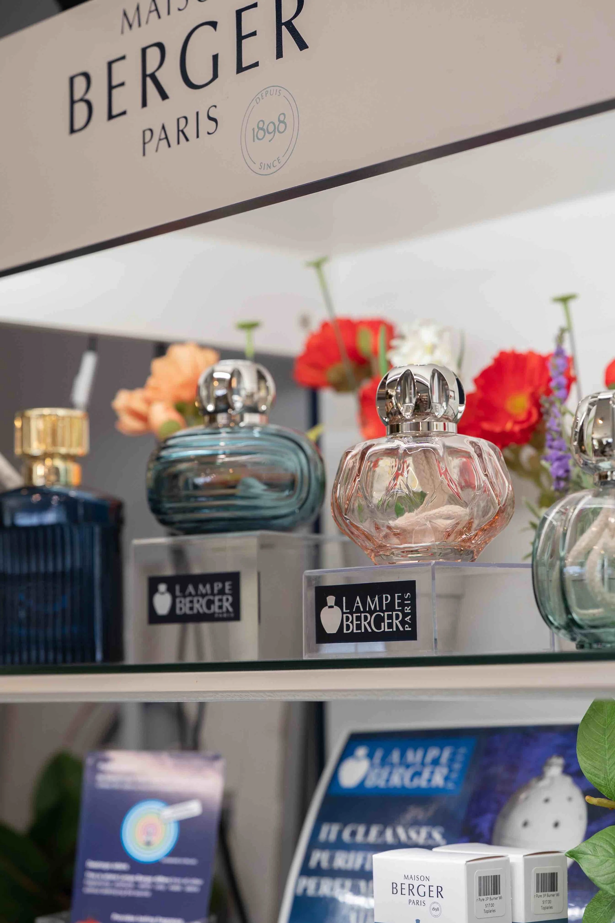 Display of perfume bottles and floral decorations on a shelf, with a sign featuring the 'Lampe Berger Paris' logo and branding.