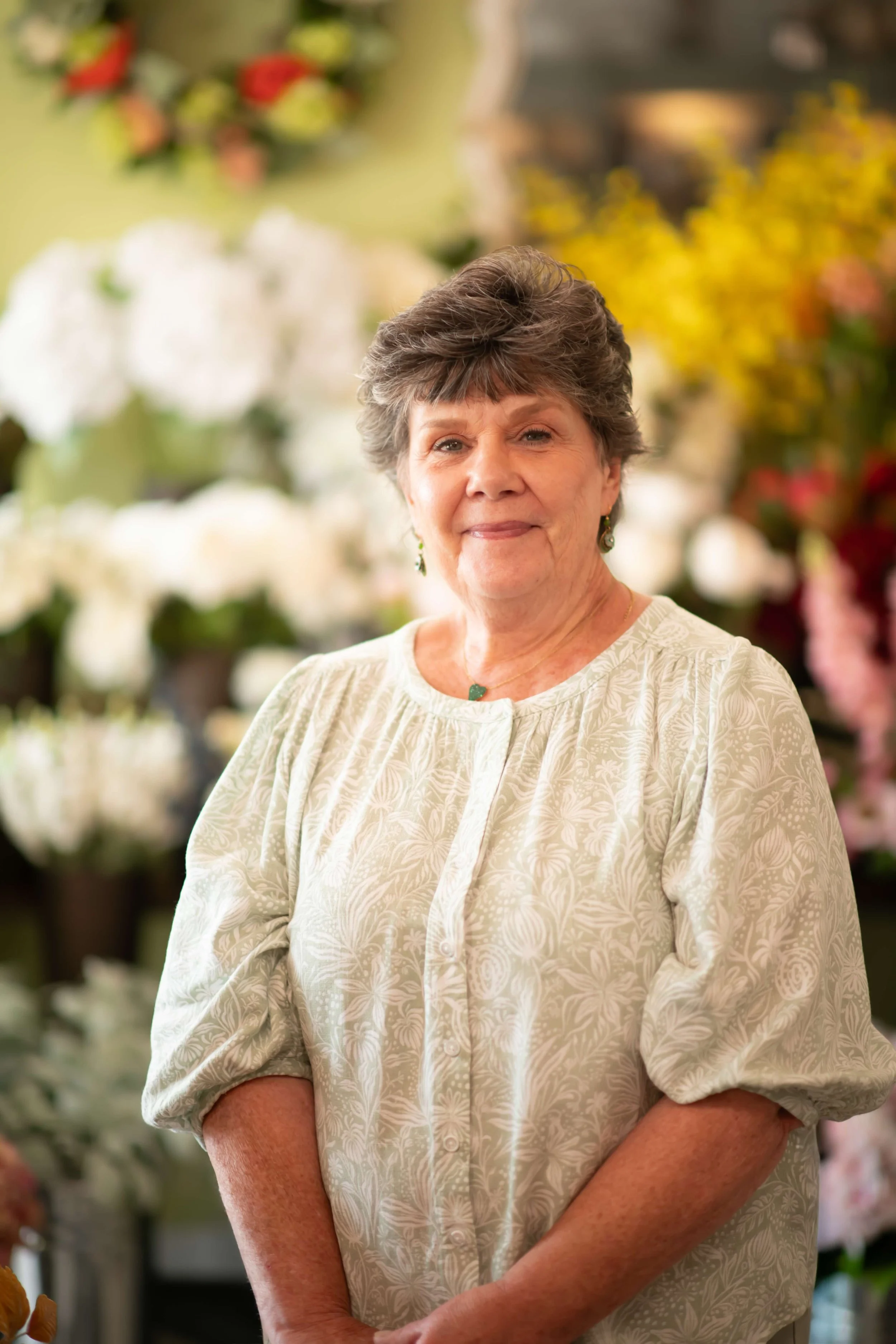 An elderly woman with short gray hair smiling at the camera, wearing a patterned light beige blouse with rolled-up sleeves, standing in a flower shop with blurred colorful flowers in the background.