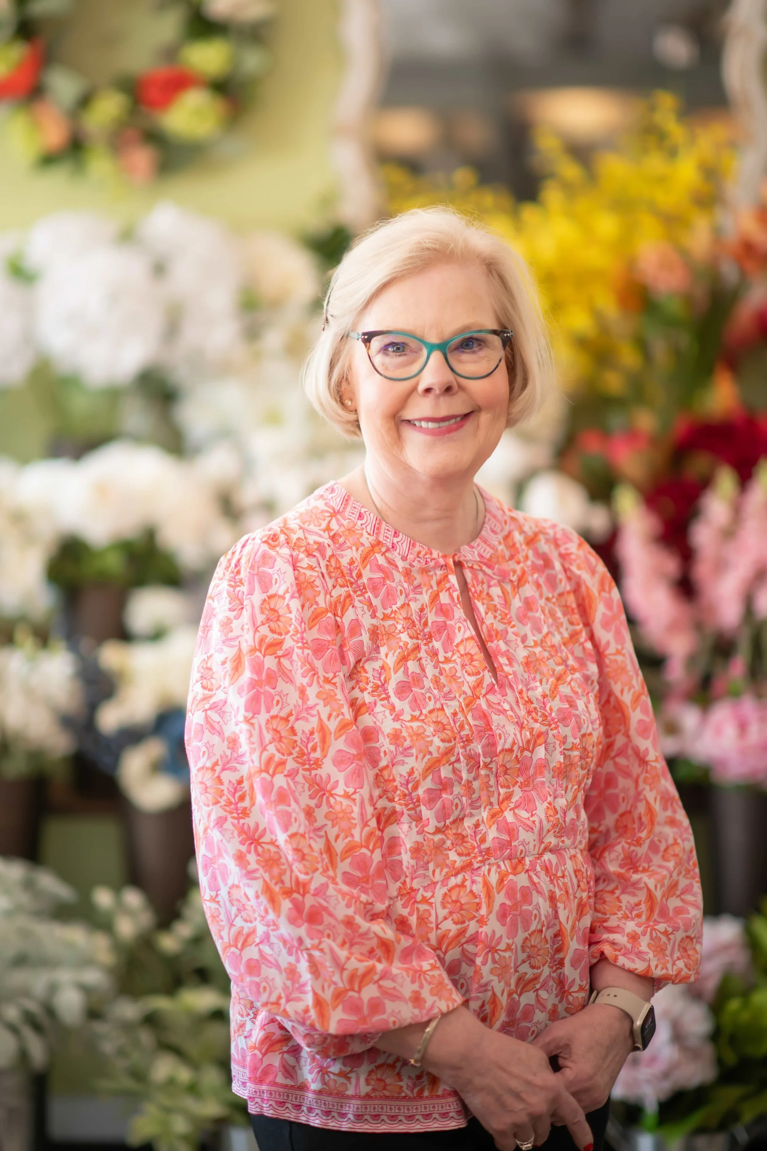 A smiling elderly woman with white hair and glasses standing in a flower shop surrounded by various colorful flowers.