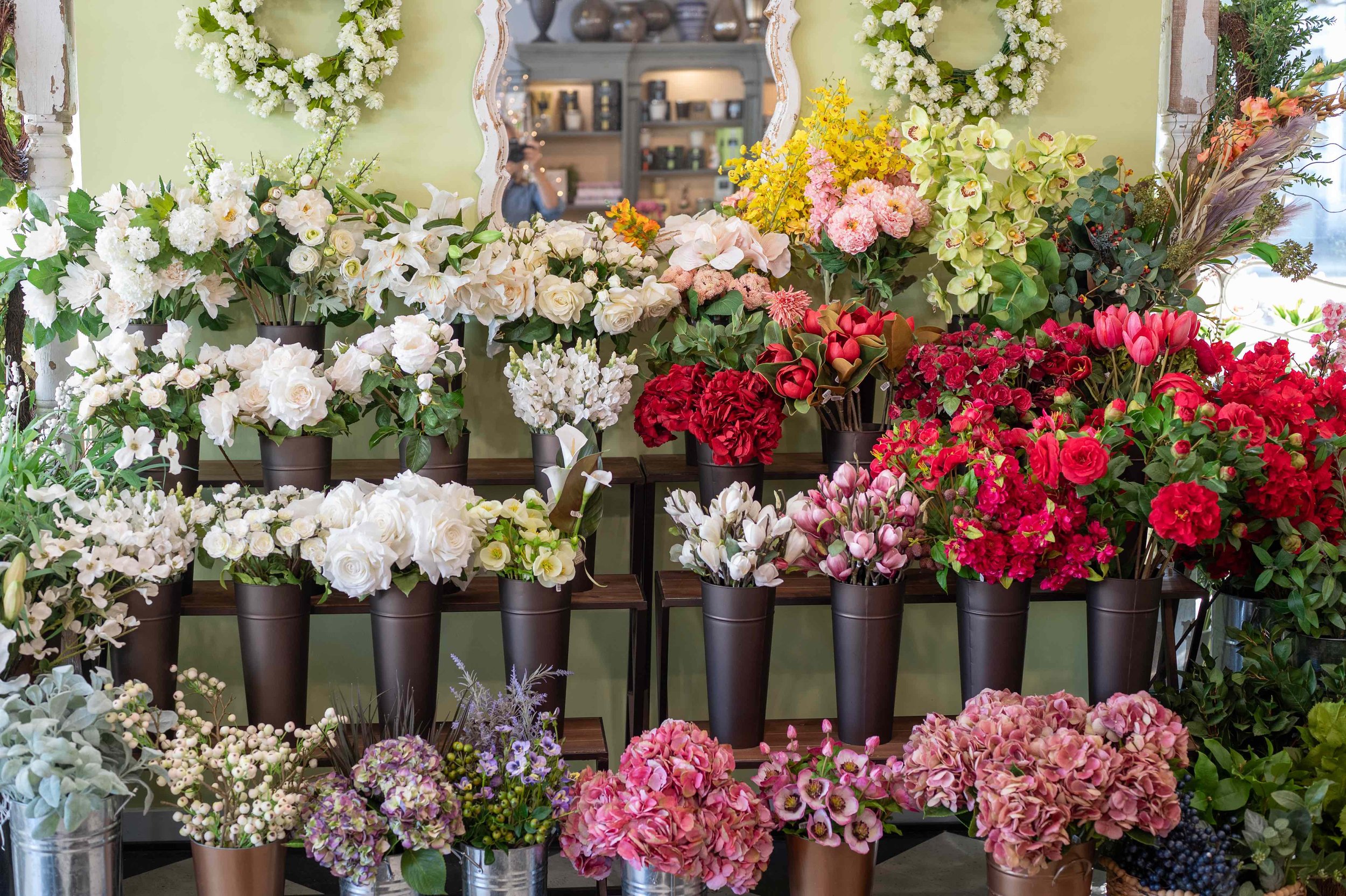 A display of various colorful flowers in black pots inside a floral shop or store, with a mirror in the background reflecting other flower arrangements and shop shelves.