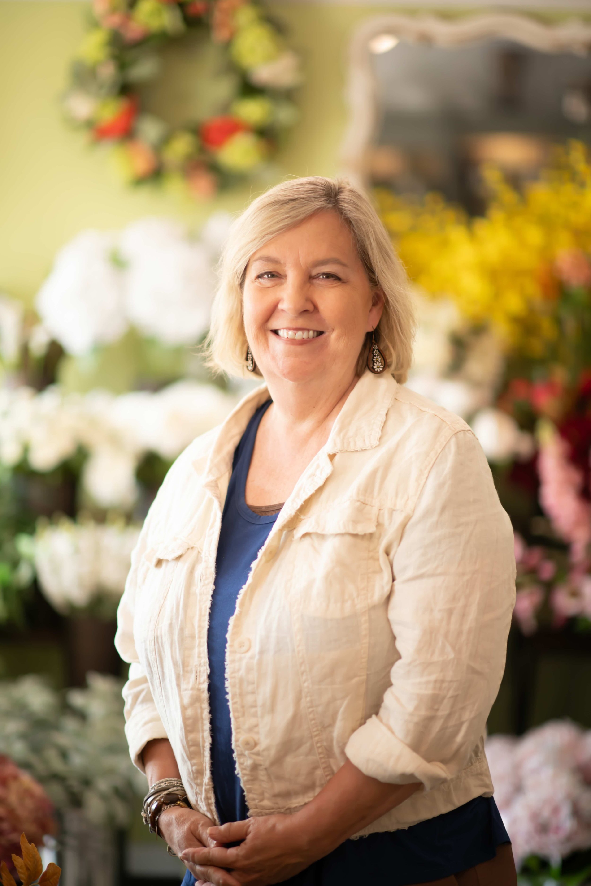 A smiling middle-aged woman with light blonde hair and earrings, standing in front of a display of colorful flowers at a flower shop or market.