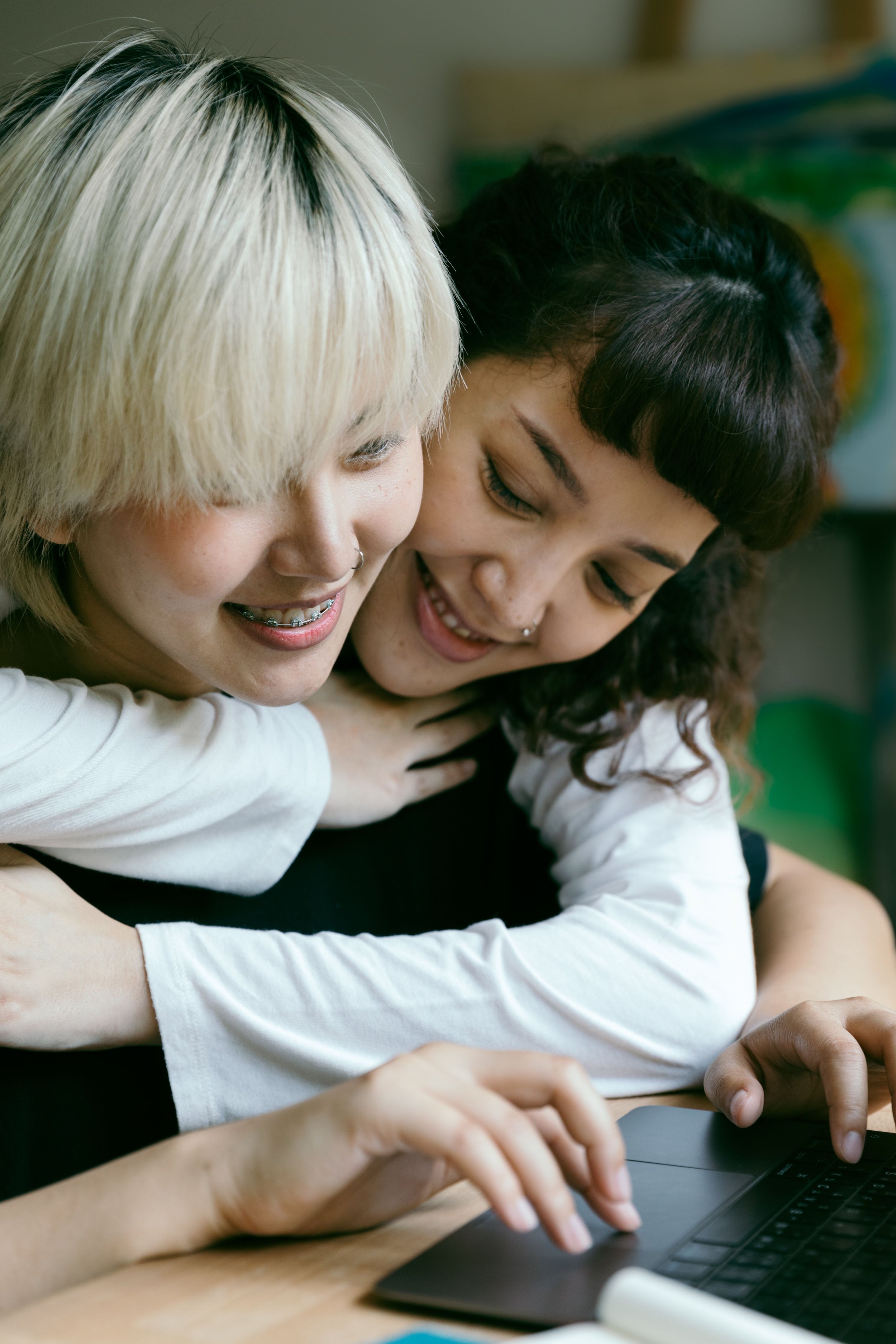 Two women smiling and hugging while using a laptop at a desk. Queer, LGBTQ+ affirmative couples therapy.