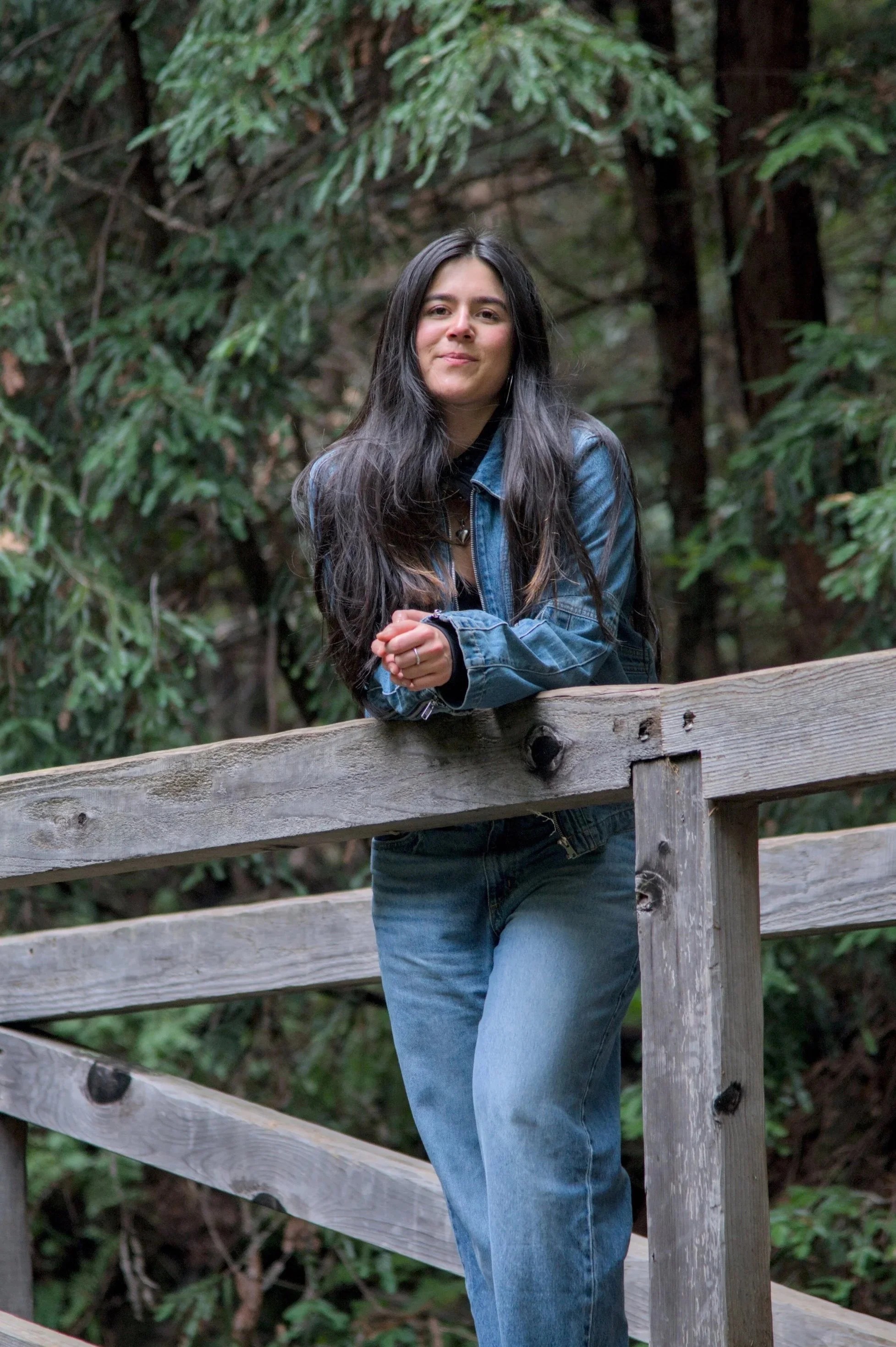 A woman with long dark hair, wearing a denim jacket and jeans, standing outdoors behind a wooden fence with greenery and trees in the background.

New York Therapist standing outdoors.