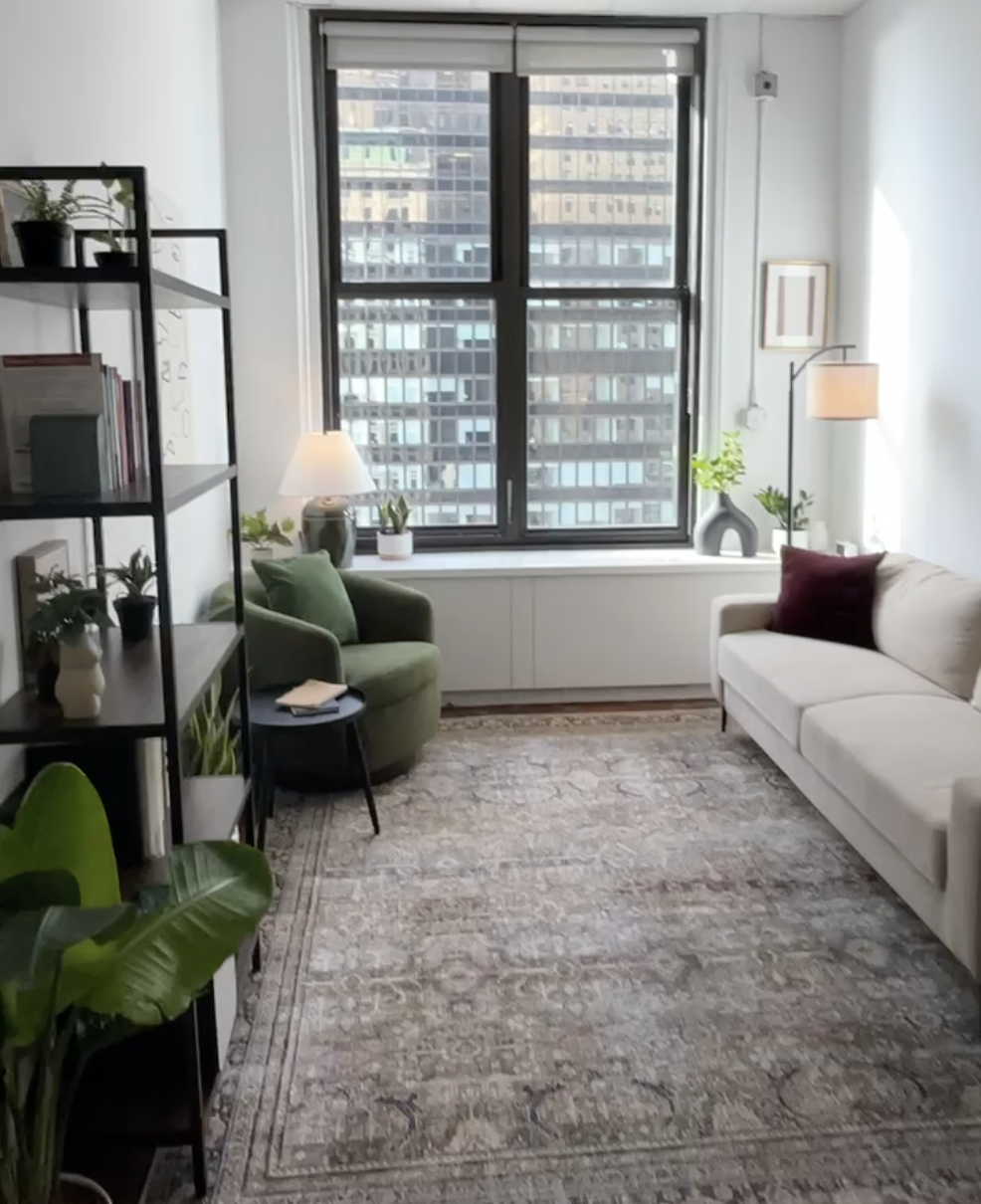 A modern living room with a large window showing city buildings outside. The room is decorated with a beige sofa with a dark red pillow, a green armchair, a black shelf with plants and books, a side table with a white lamp, and a standing lamp with a beige shade.