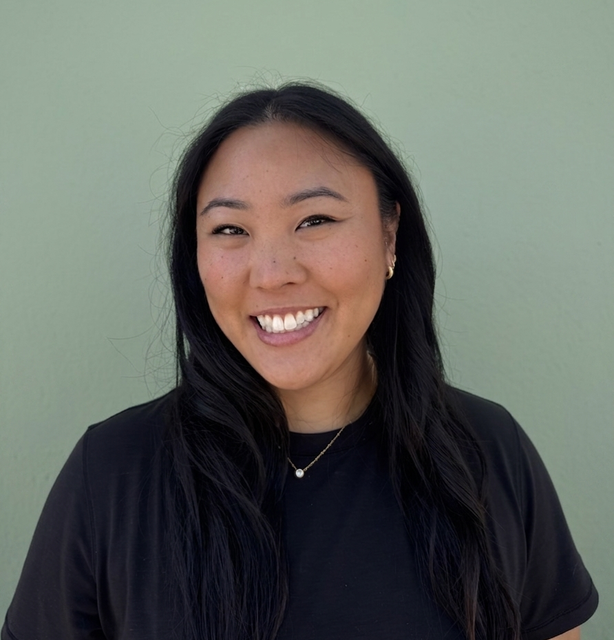 Young woman with long black hair smiling in front of a light green wall, wearing a black top and a pearl necklace.