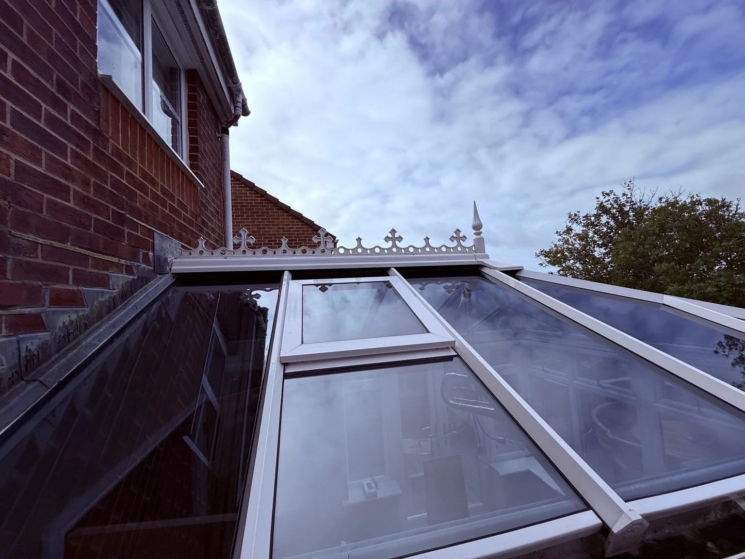 A glass conservatory roof attached to a brick house with a Victorian-style decorative finial on top and an overcast sky in the background.