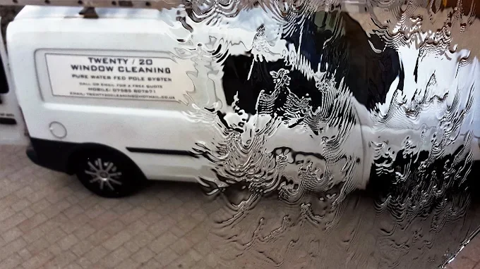 A van with a sign that reads "TWENTY / 20 WINDOW CLEANING" is seen through a window with water or soap on it, creating a distorted and wavy reflection.