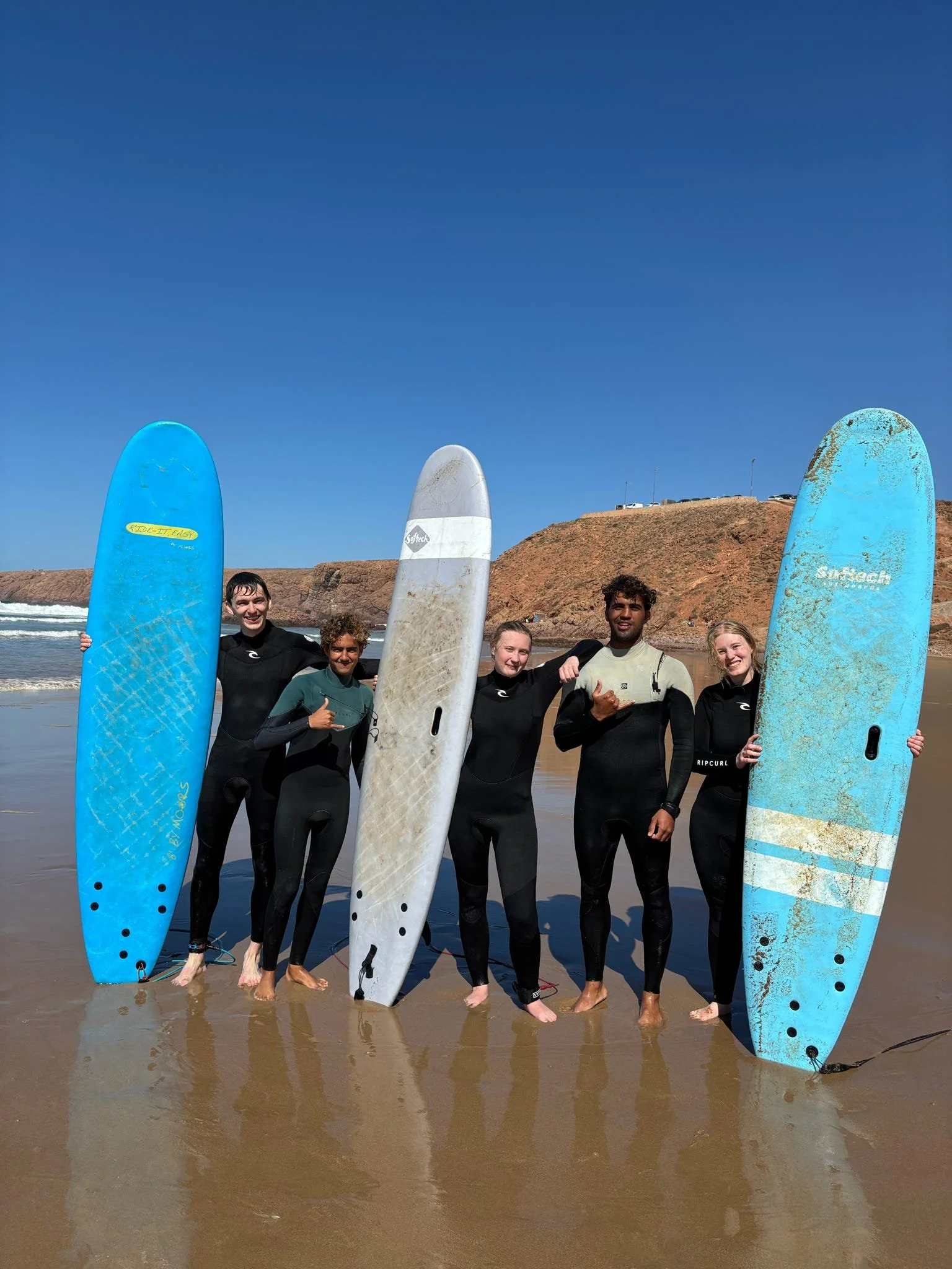 A group of six surfers standing on the wet sand at the beach, holding surfboards, with the ocean and a sandy hill in the background under a blue sky.
