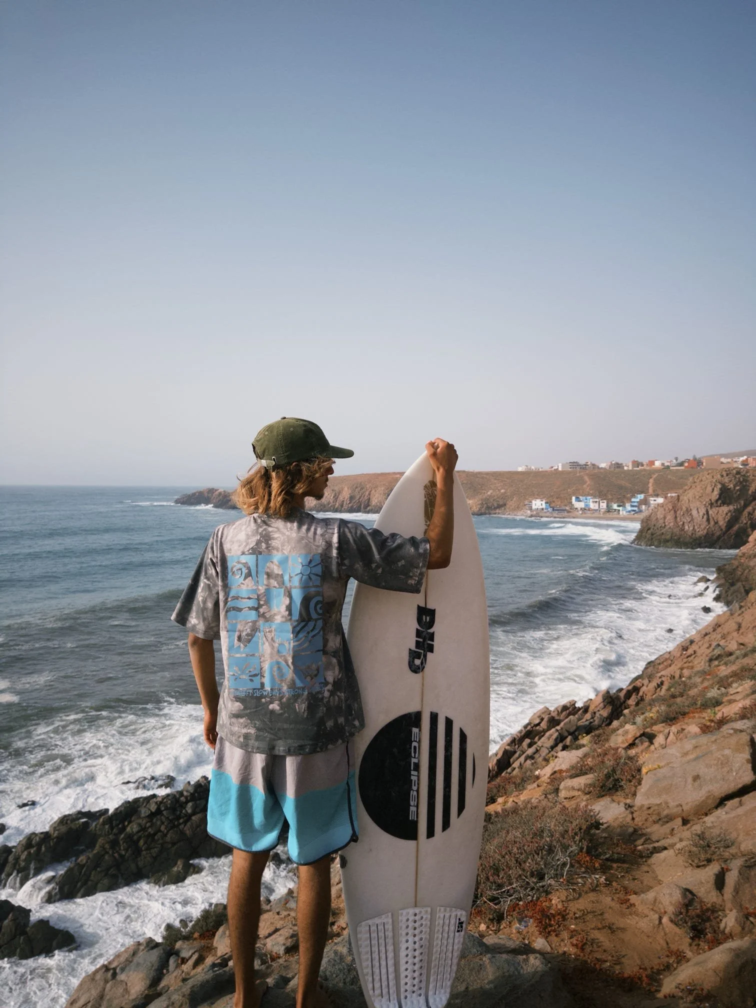 A person holding a surfboard on a rocky beach with ocean waves and cliffs in the background.