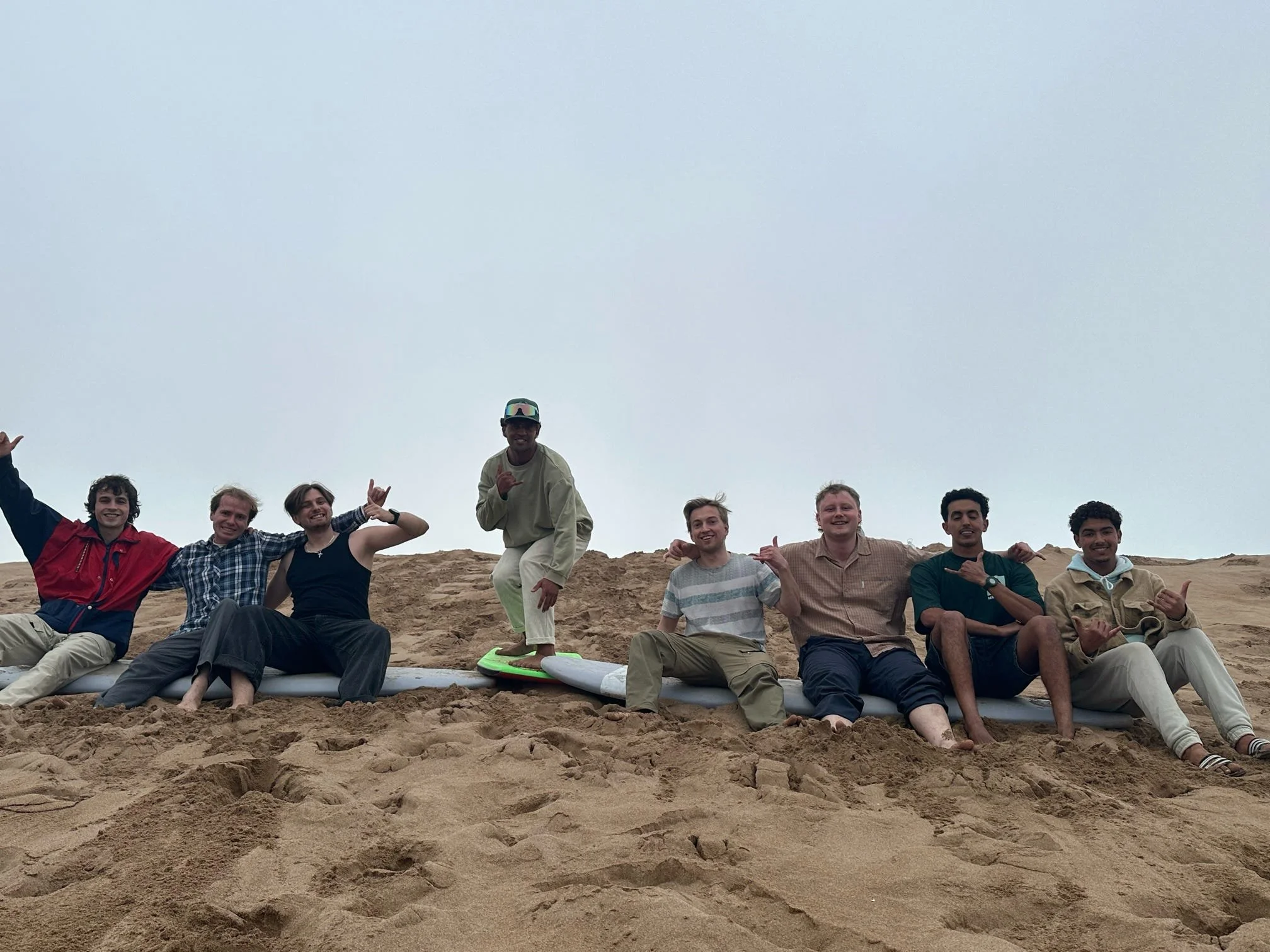 A group of people sitting and standing on a sandy beach, smiling and posing for a photo, with one person in the center kneeling on a surfboard and wearing a helmet.