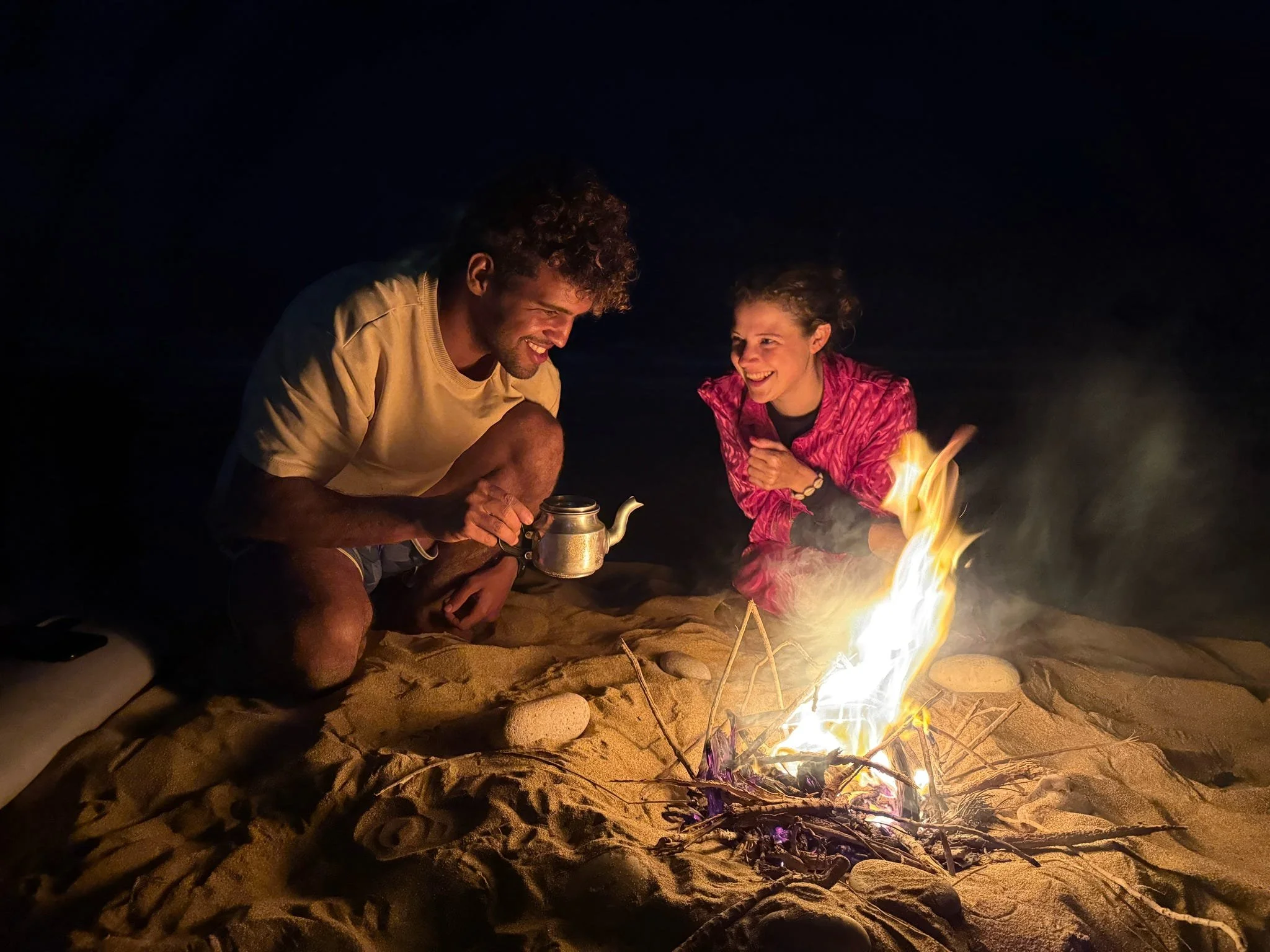 A man and a woman sitting close by a fire on the beach at night, smiling and enjoying each other's company. The man is holding a teapot, and the woman is wearing a pink jacket.