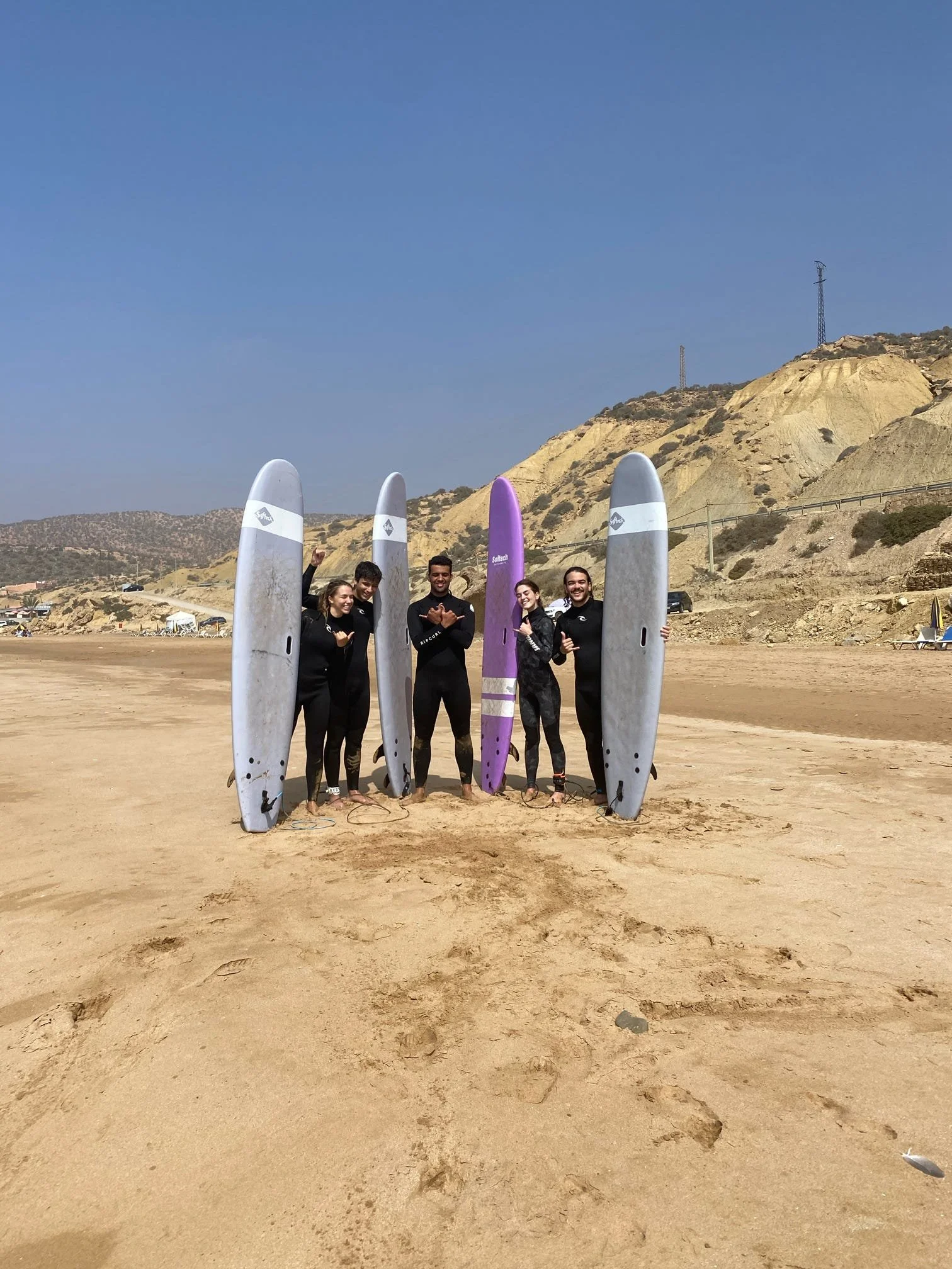 A group of five people standing on a sandy beach, each holding a surfboard, with a mountainous landscape in the background and a clear blue sky.