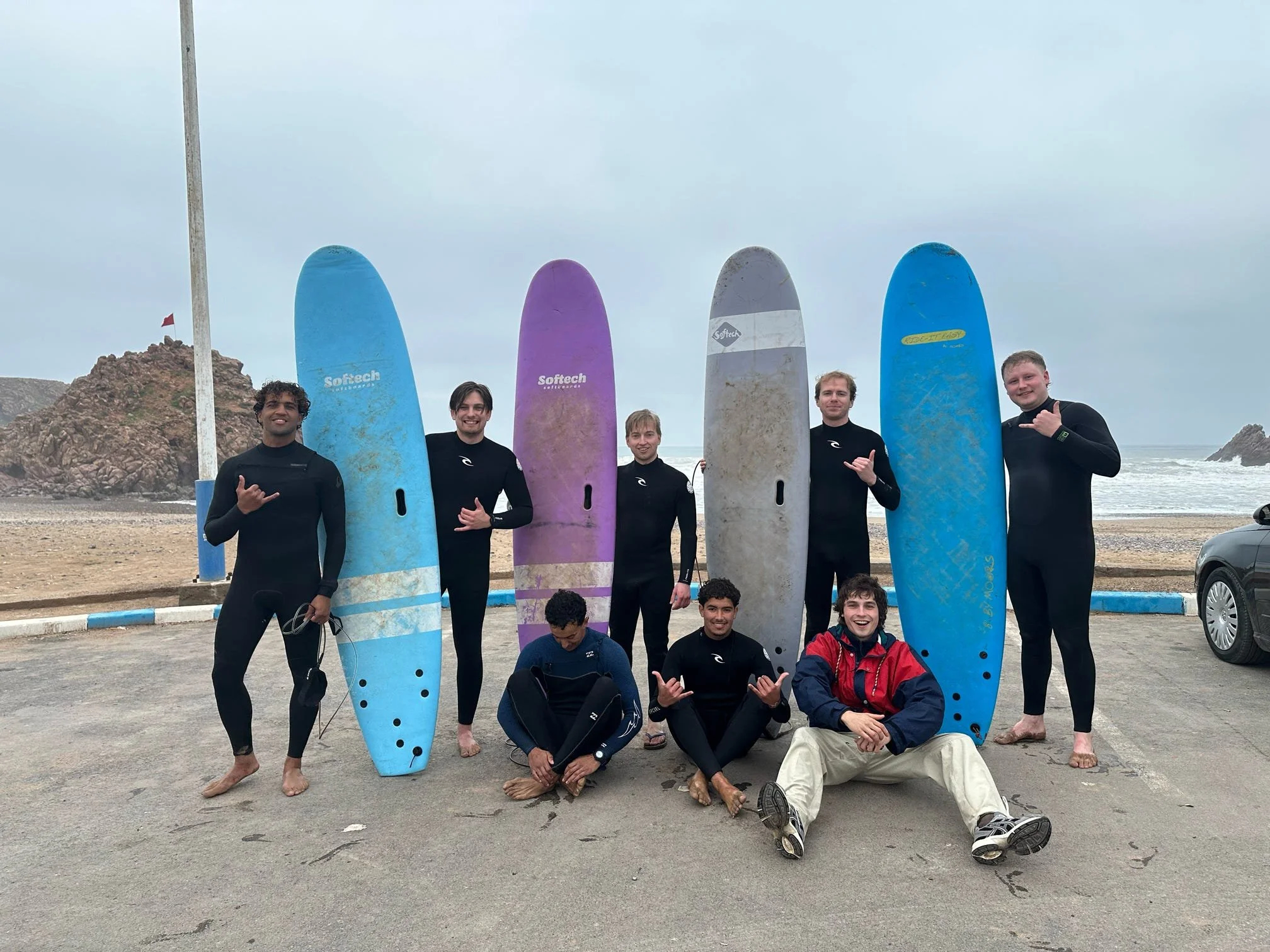 Group of young surfers holding colorful surfboards on the beach, with a rocky hill and cloudy sky in the background.