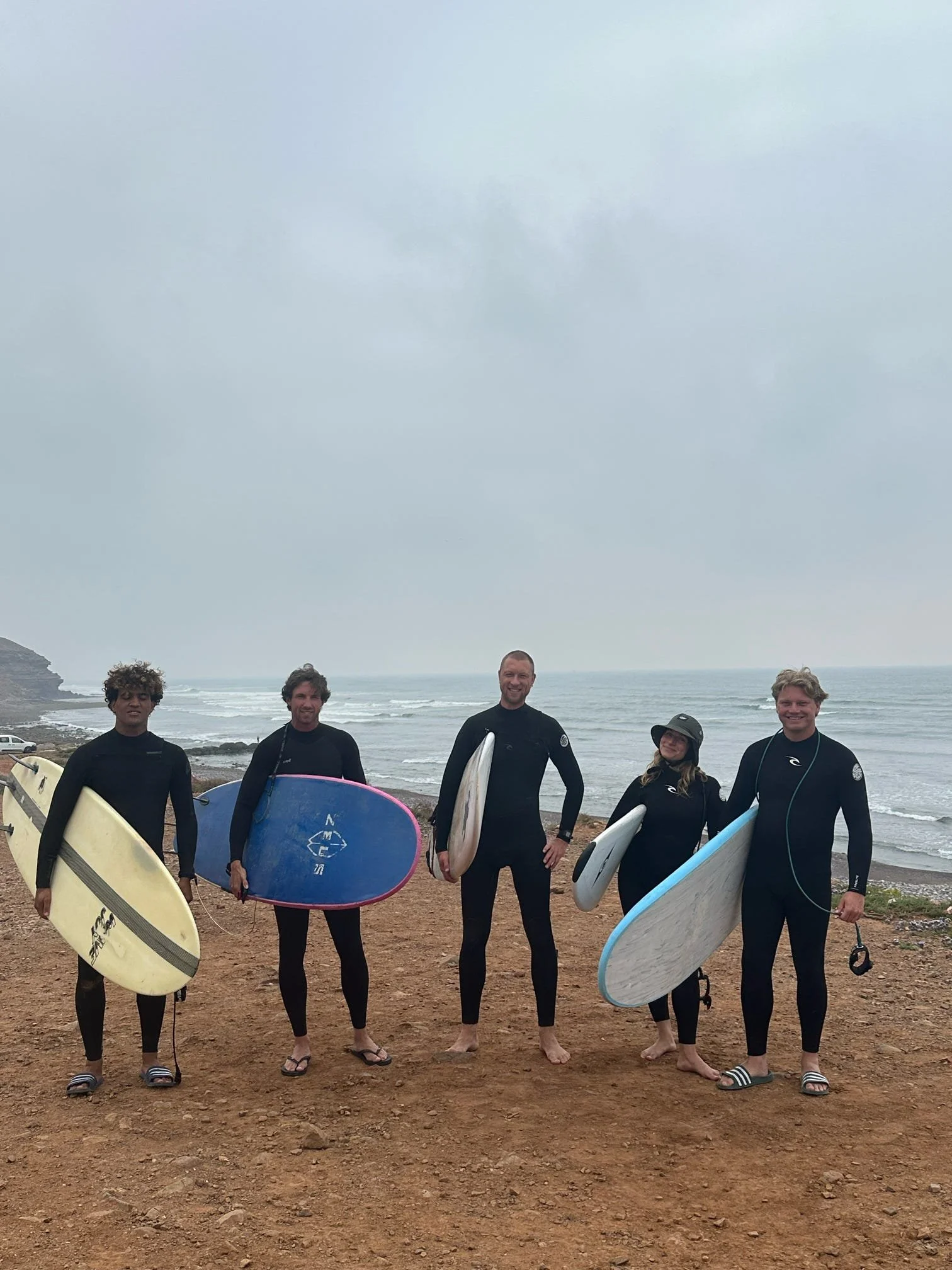 Group of five surfers standing on a beach holding surfboards, with the ocean and cloudy sky in the background.