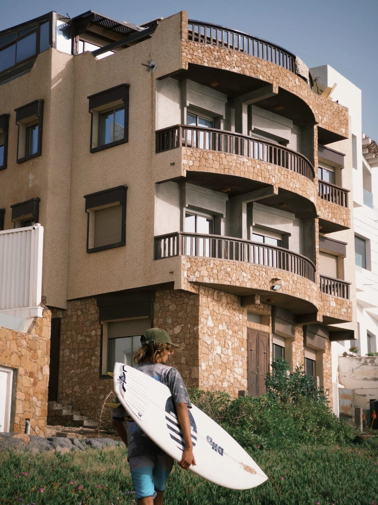 A person with long hair, wearing a cap, a dark shirt, and cargo shorts, carrying a surfboard and walking on a grassy area with small pink flowers in front of a multi-story building with stone and stucco exterior and curved balconies.