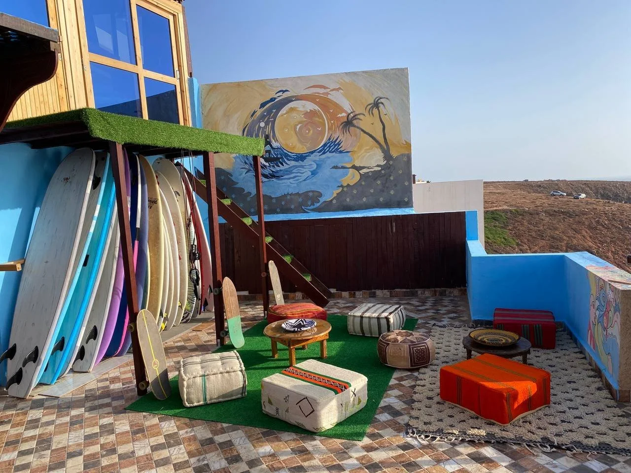 Outdoor patio with surfboards, cushions, and small tables against a blue and yellow wall. A mural of a wave and moon on the wall, with clear sky and hills in the background.