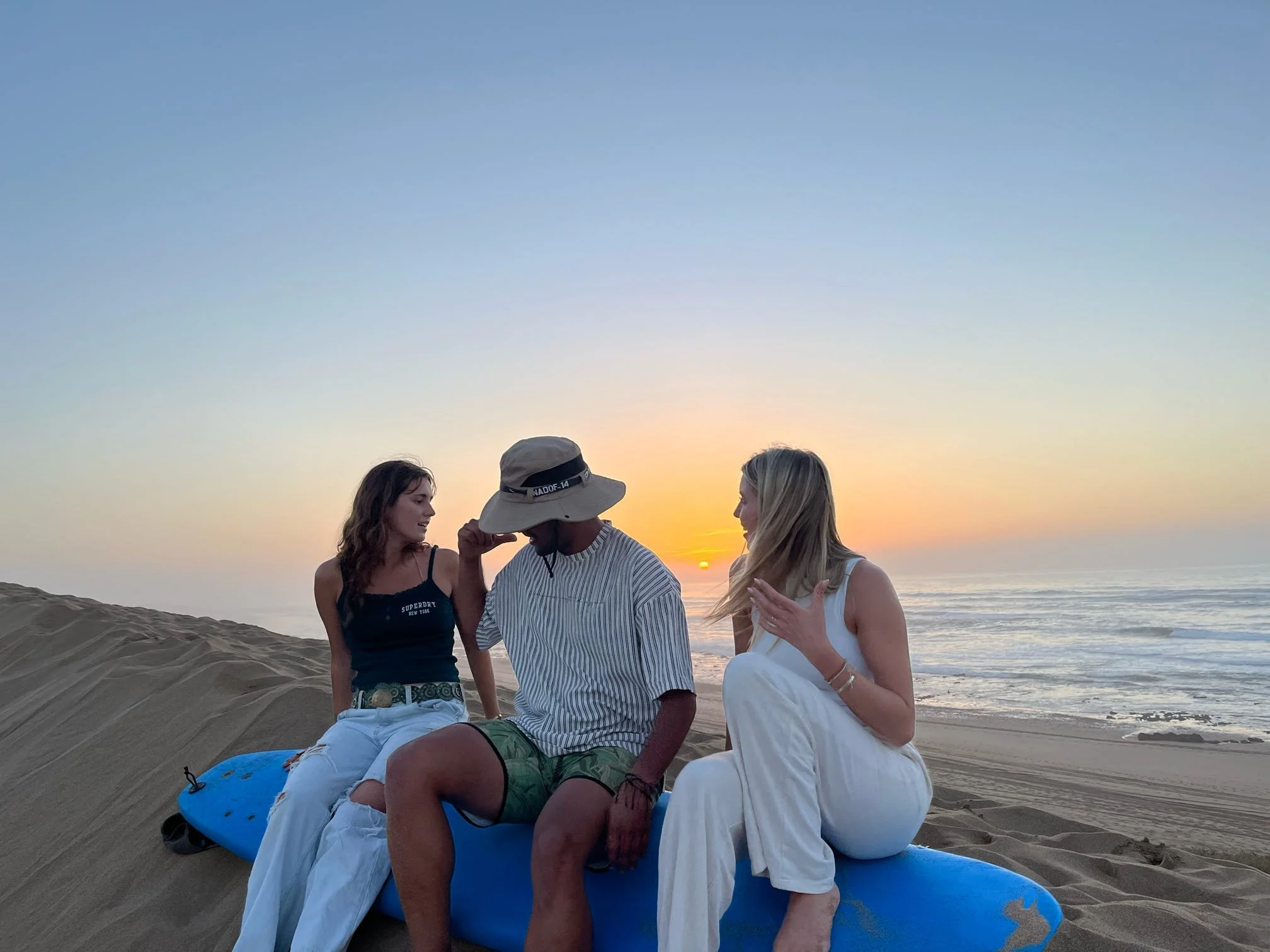 Four people sitting on the sand at the beach during sunset, with the ocean in the background.