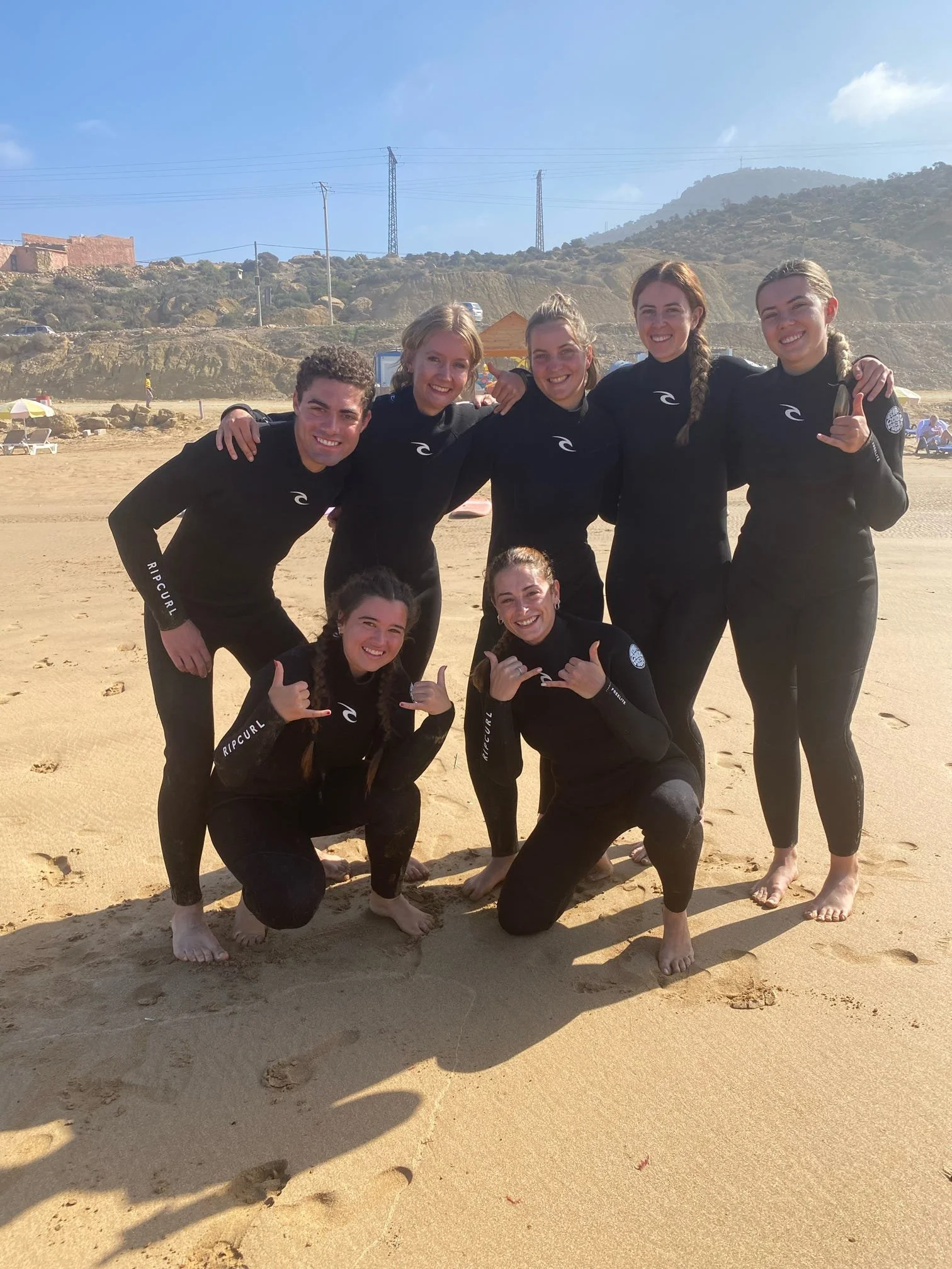 Group of six young people in wetsuits posing together on a sandy beach, smiling and making hand gestures, with mountains and electrical towers in the background.
