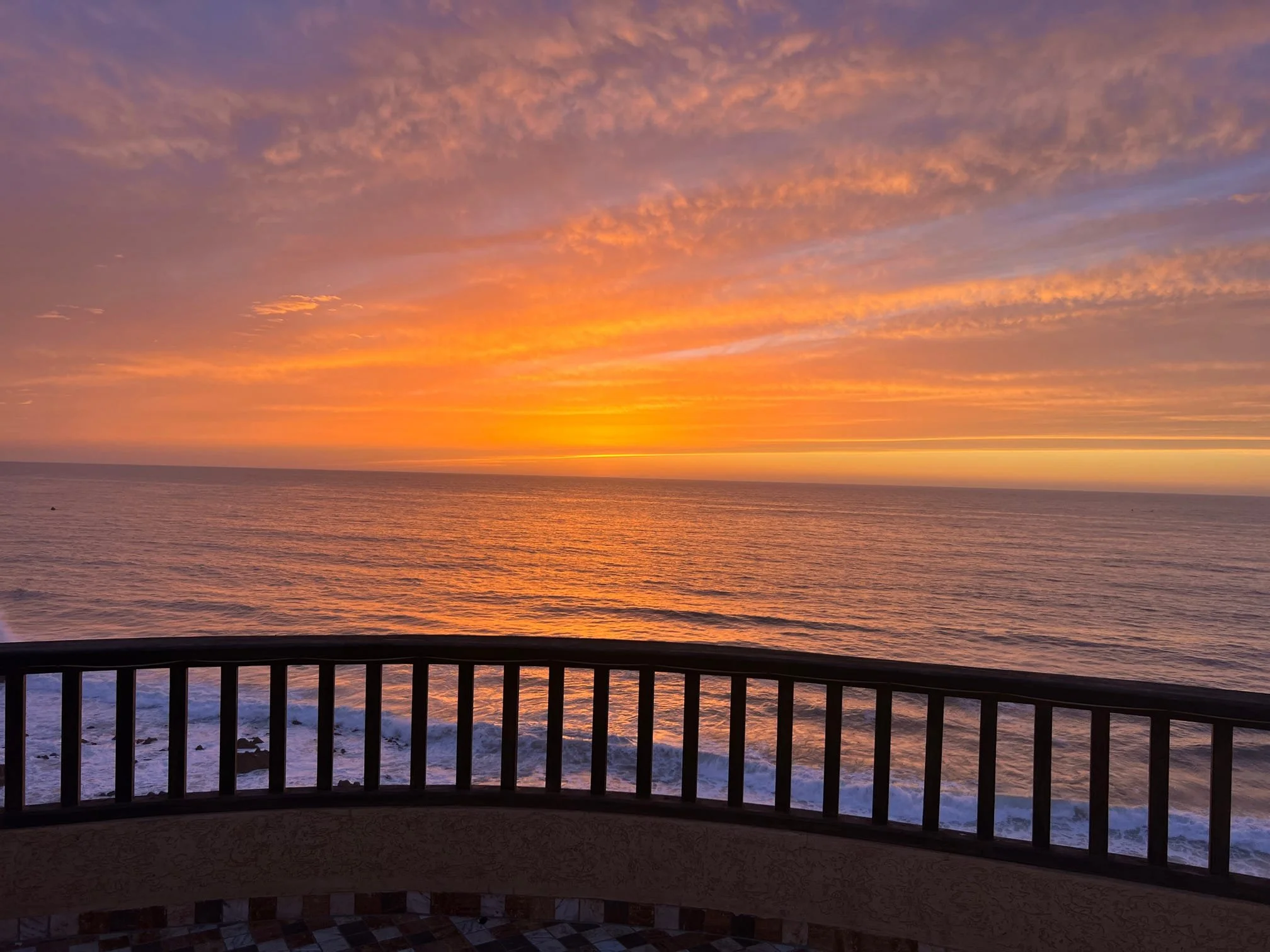 Sunset over the ocean viewed from a balcony with a black railing.