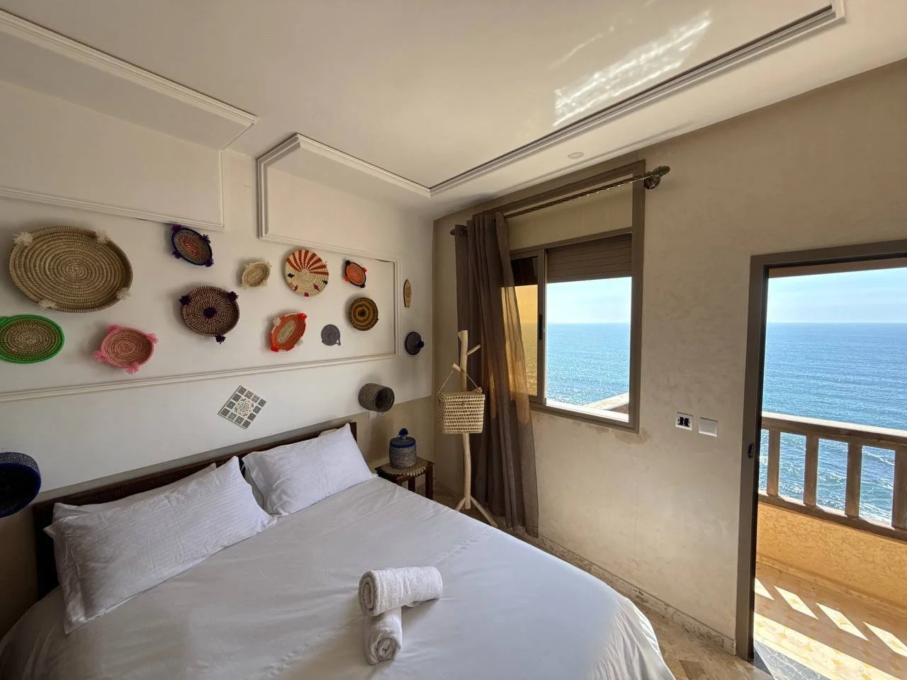 Hotel bedroom with white bedding, decorative woven wall art, a window with a view of the ocean, and a small balcony.