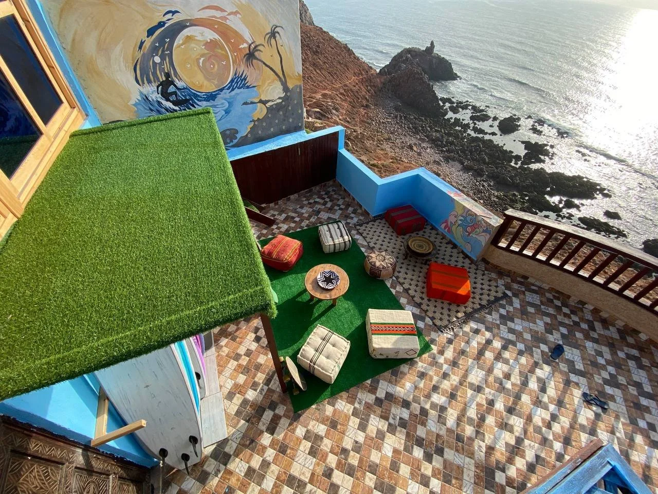 Beachside outdoor patio with colorful decor, a small table, cushions, and a mural on the wall, overlooking rocky shoreline and ocean.