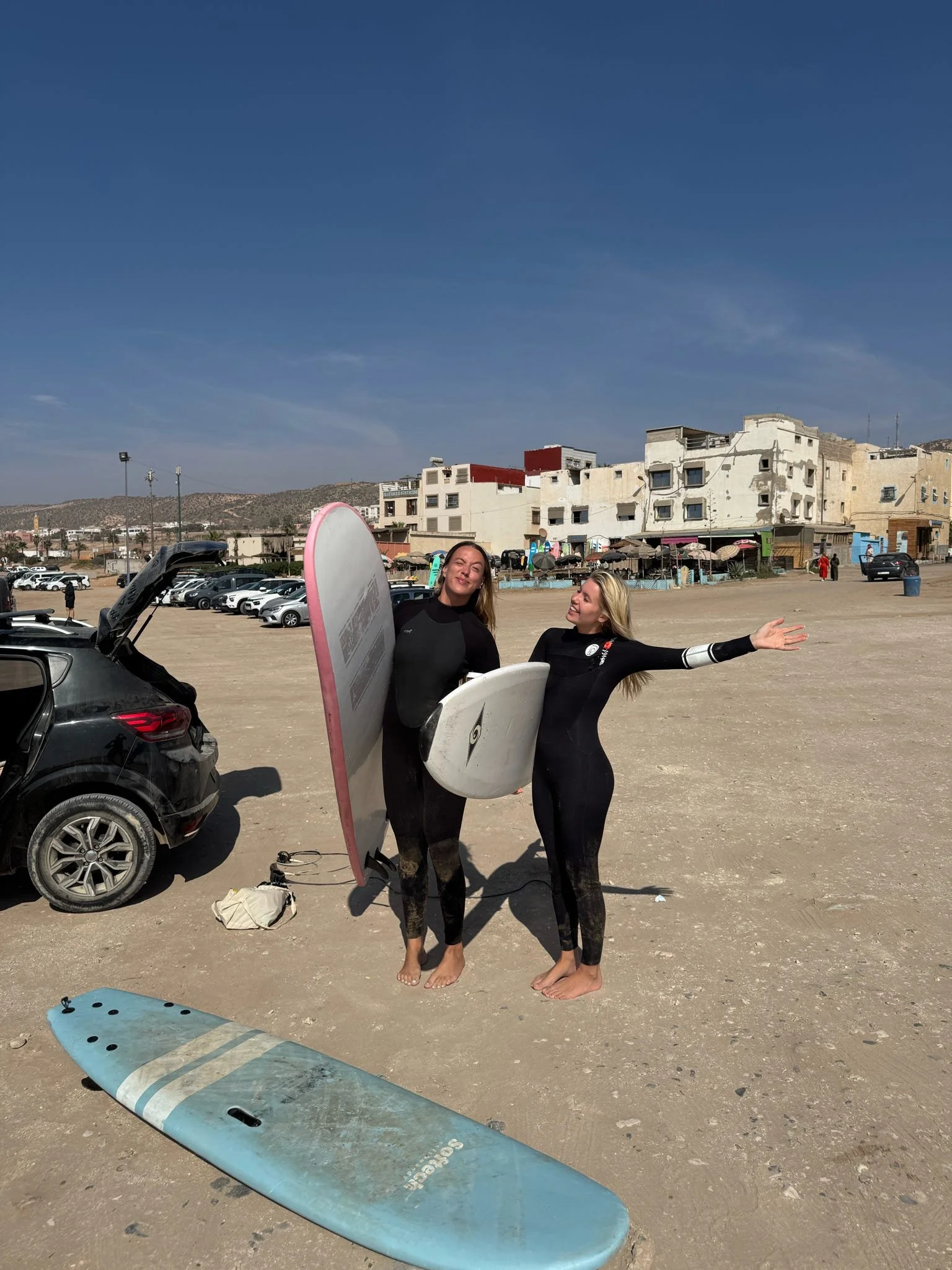Two women in wetsuits holding surfboards, standing on a sandy beach with parked cars, a building, and a hill in the background.