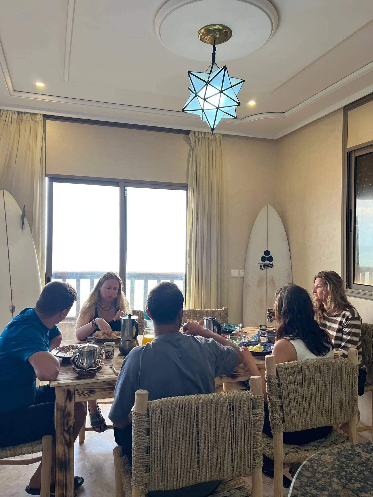 A group of people are sitting at a wooden dining table enjoying a meal together in a bright room with large windows, cream curtains, and a decorative ceiling light.