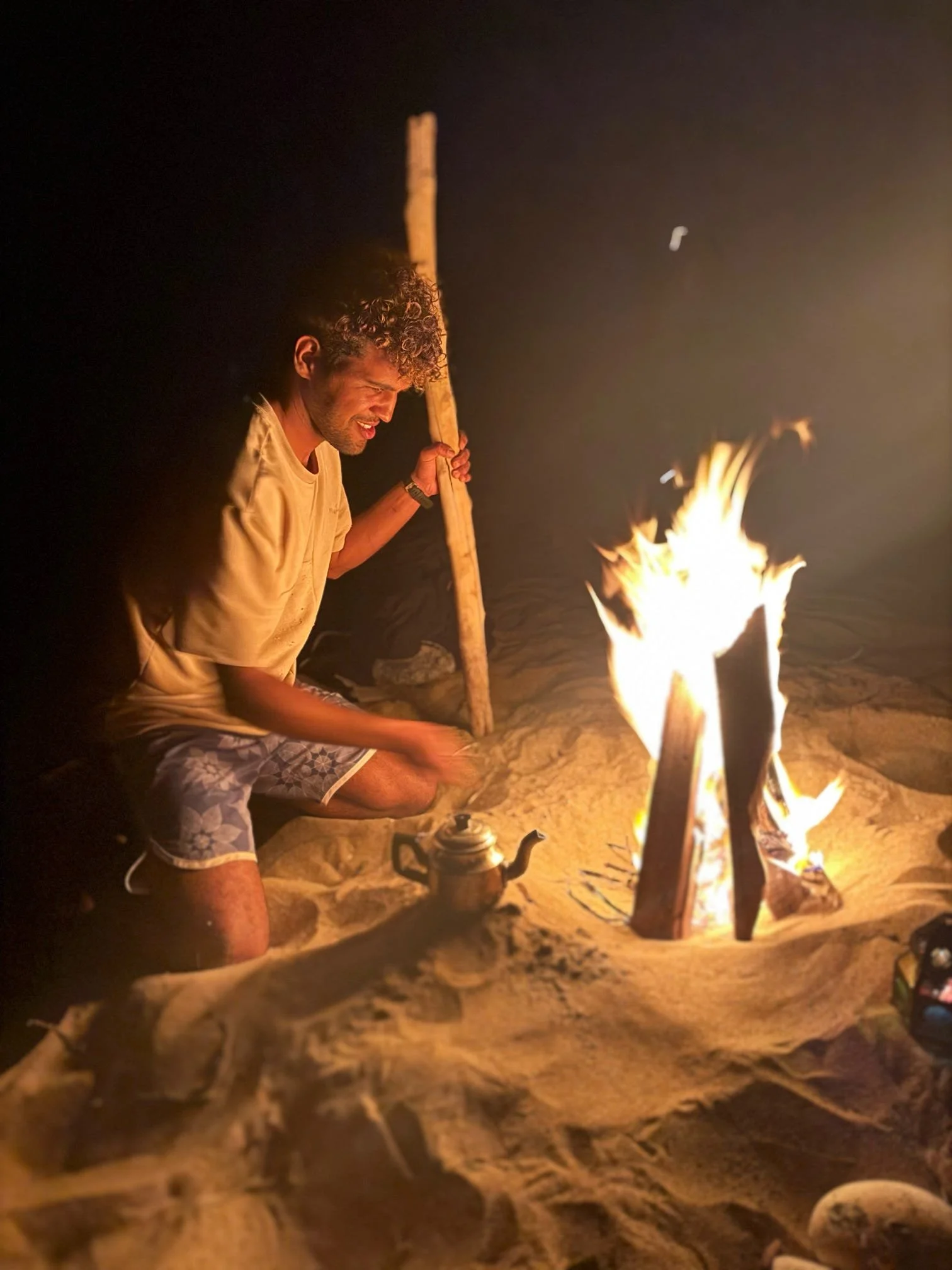 A young man with curly hair, wearing a yellow shirt and shorts, sitting on the sand at night by a campfire, holding a long stick, with a kettle nearby.