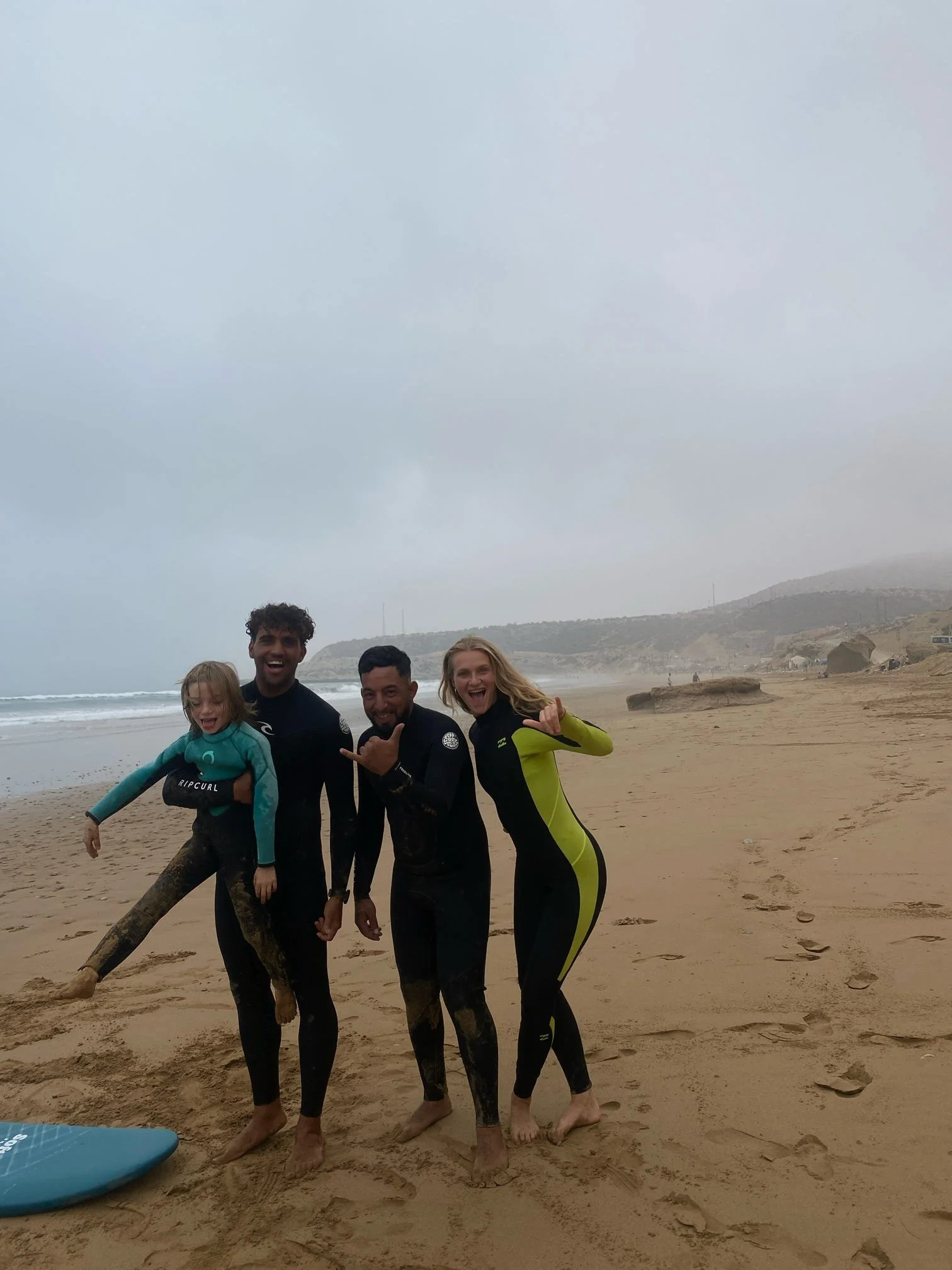 A group of five people, including a child, smiling and posing on a sandy beach with the ocean and hills in the background. They are dressed in wetsuits, indicating they are engaging in water sports or swimming.