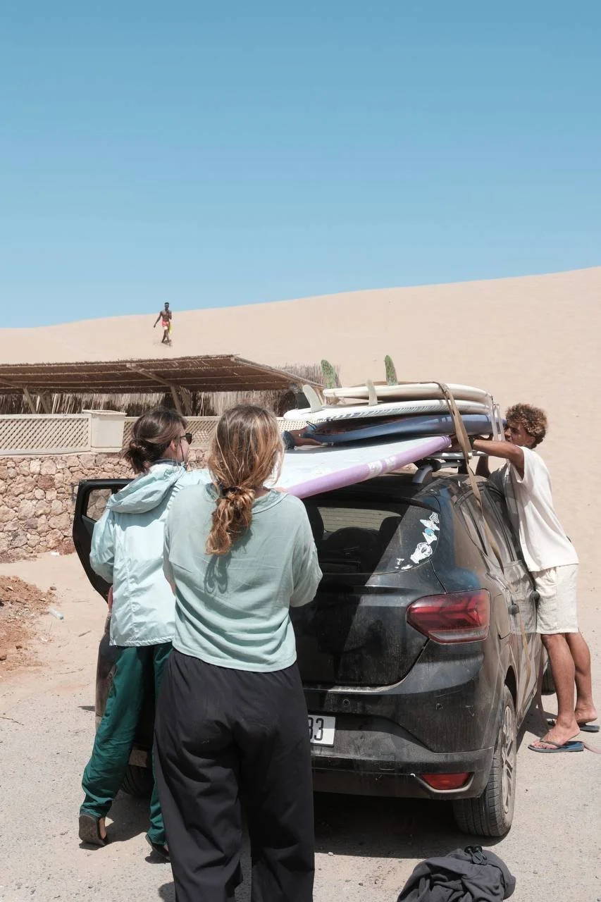 Four people preparing surfboards on the roof of a black car near a sand dune with one person walking on the sand in the background during daytime.