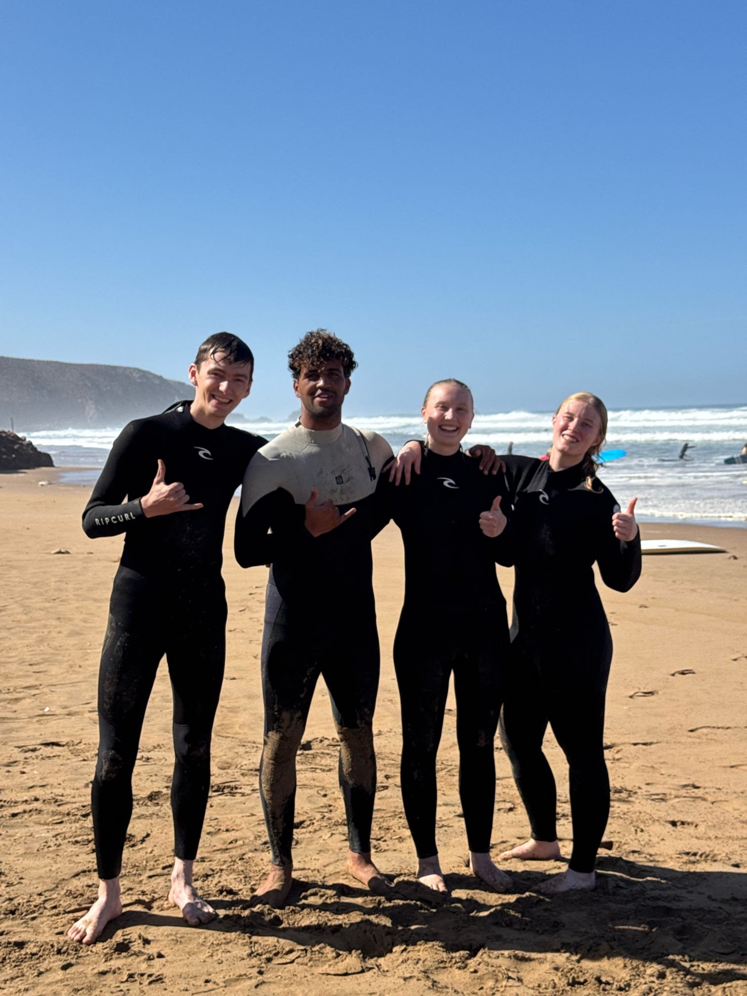 Four people in wetsuits standing on a sandy beach, smiling and giving thumbs-up gestures, with the ocean and cliffs in the background on a sunny day.