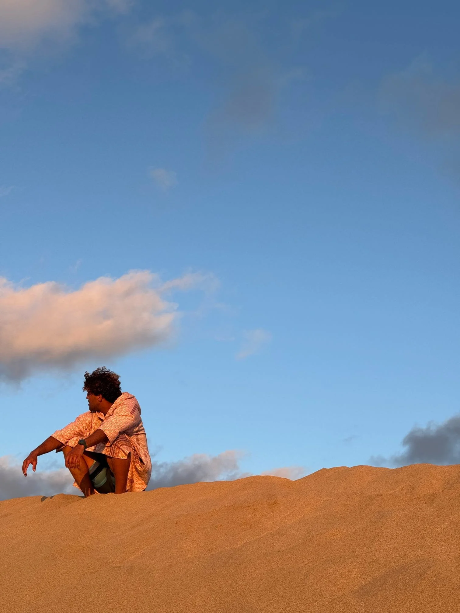 A person with curly hair and a patterned shirt leaning on a sandy terrain outdoors, with a blue sky and some clouds in the background.