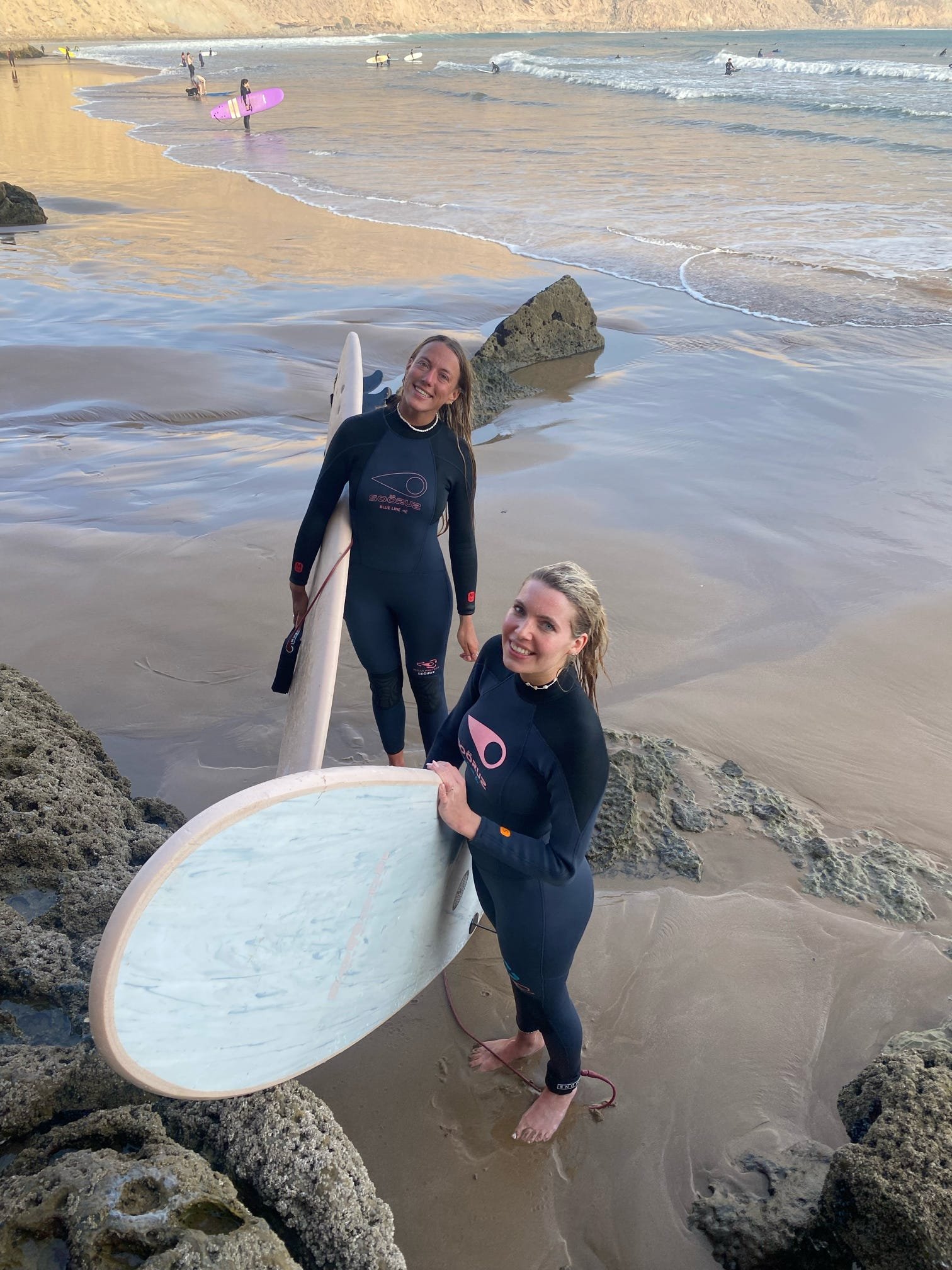 Two women in wetsuits standing on a sandy beach near rocks, holding surfboards. In the background, people are surfing and preparing to surf in the ocean, with some standing on their surfboards and others riding waves.