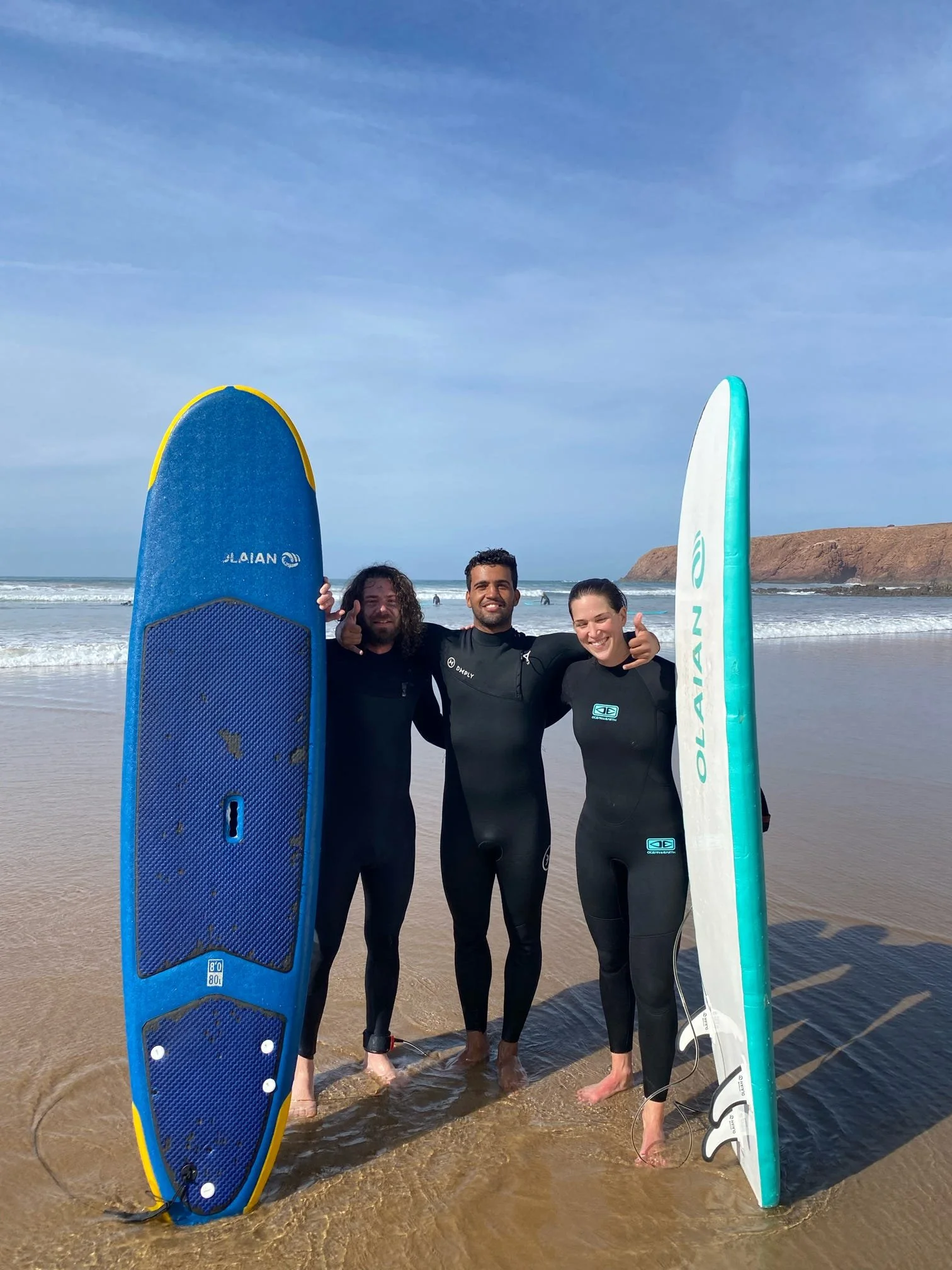 Three surfers in wetsuits standing at the edge of the ocean, holding surfboards. The sky is blue with some clouds, and there is a coastline with cliffs in the background.