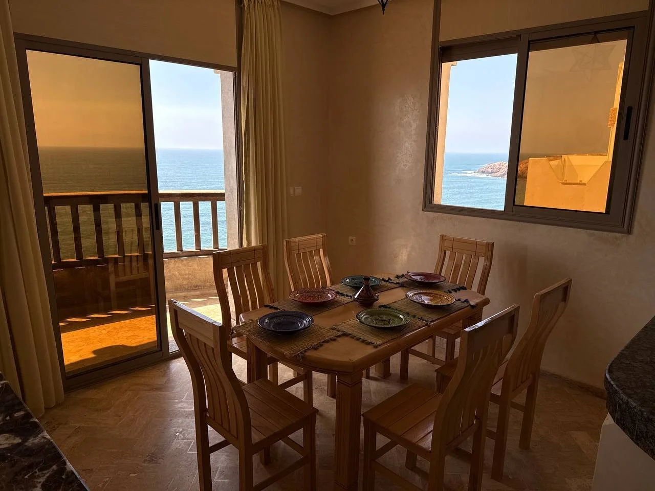 Dining area with a wooden table and six chairs, set with plates and placemats. Large windows and a sliding door lead to a balcony overlooking the ocean, with clear skies and a small island in the distance.