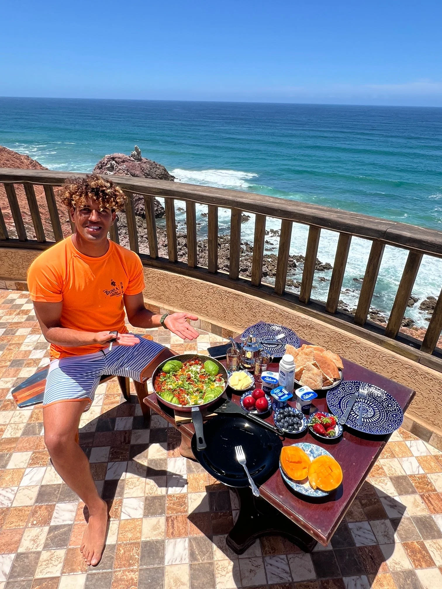 A man in an orange shirt and striped shorts is sitting outdoors with a view of the ocean, gesturing towards a table with a variety of colorful food.