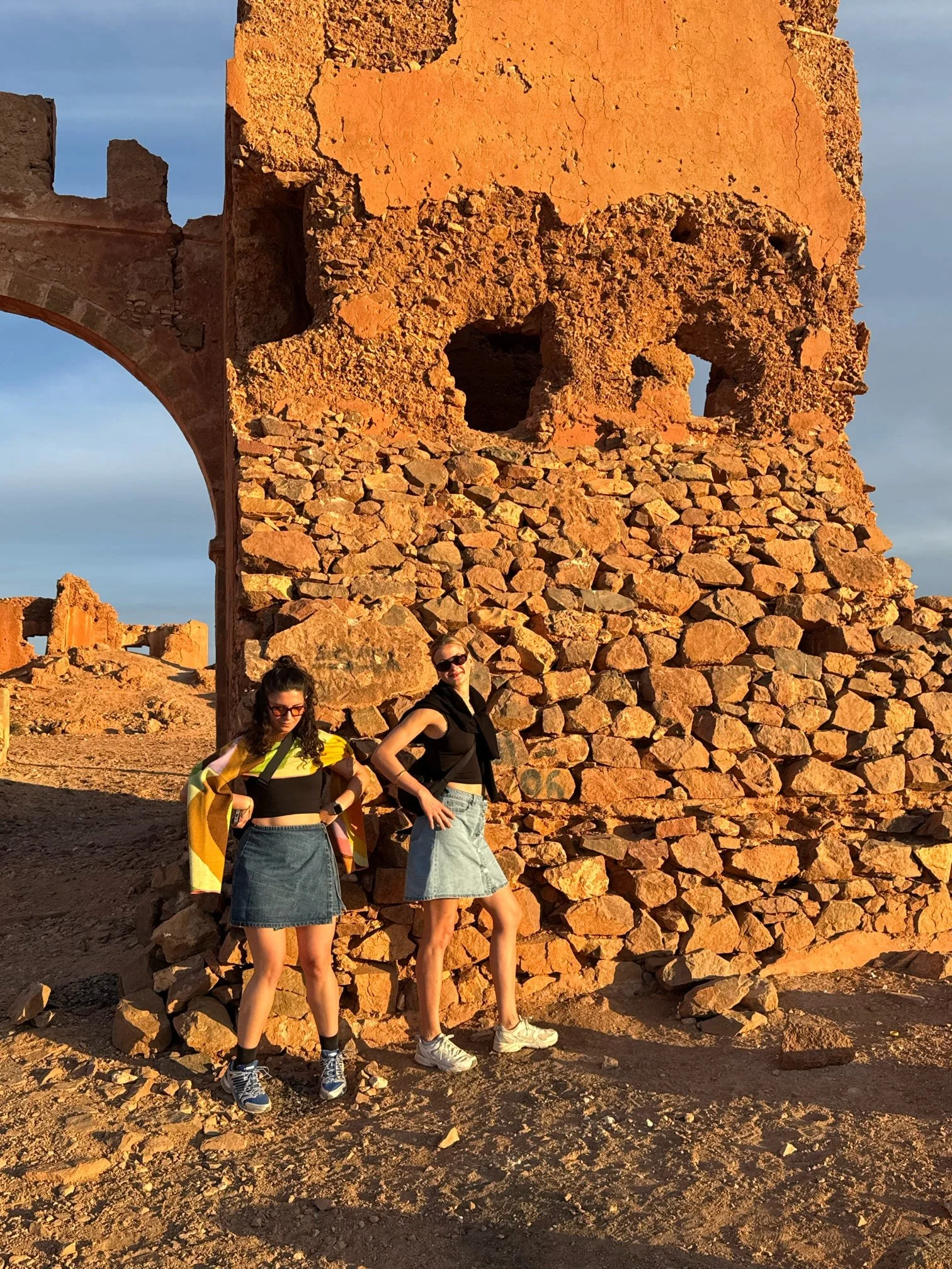 Two young women stand in front of ancient, weathered stone ruins during sunset, with rocky ground and a clear sky background.