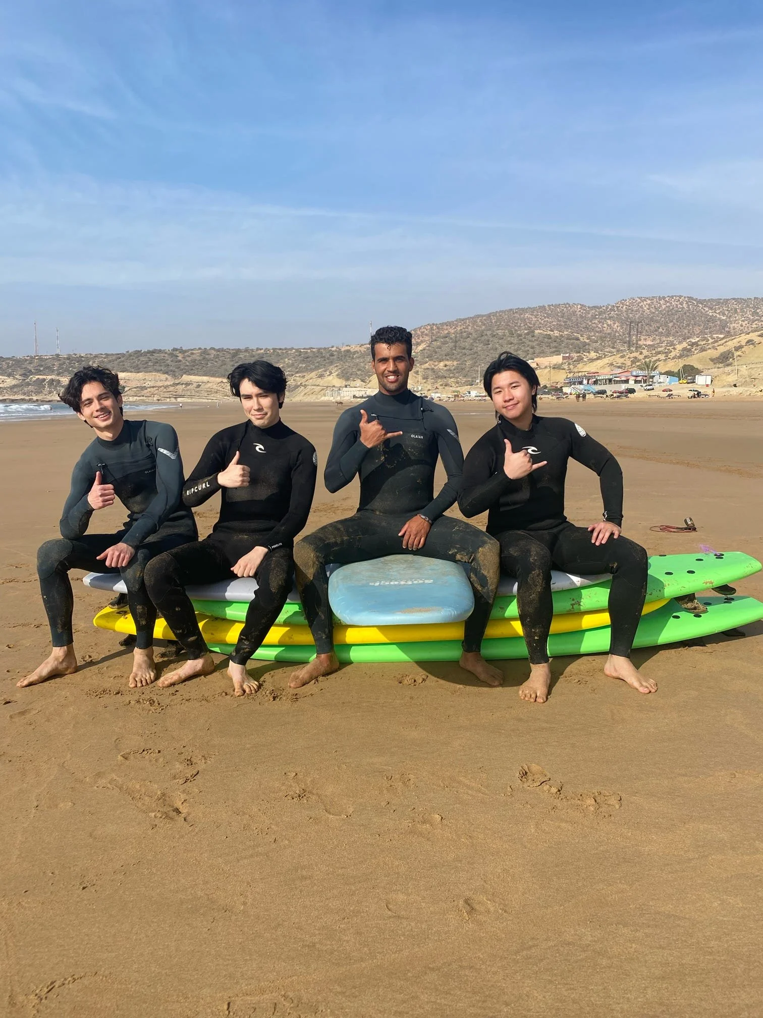 Four men in wetsuits sitting on surfboards on a sandy beach, smiling and making hand gestures, with hills and buildings in the background.
