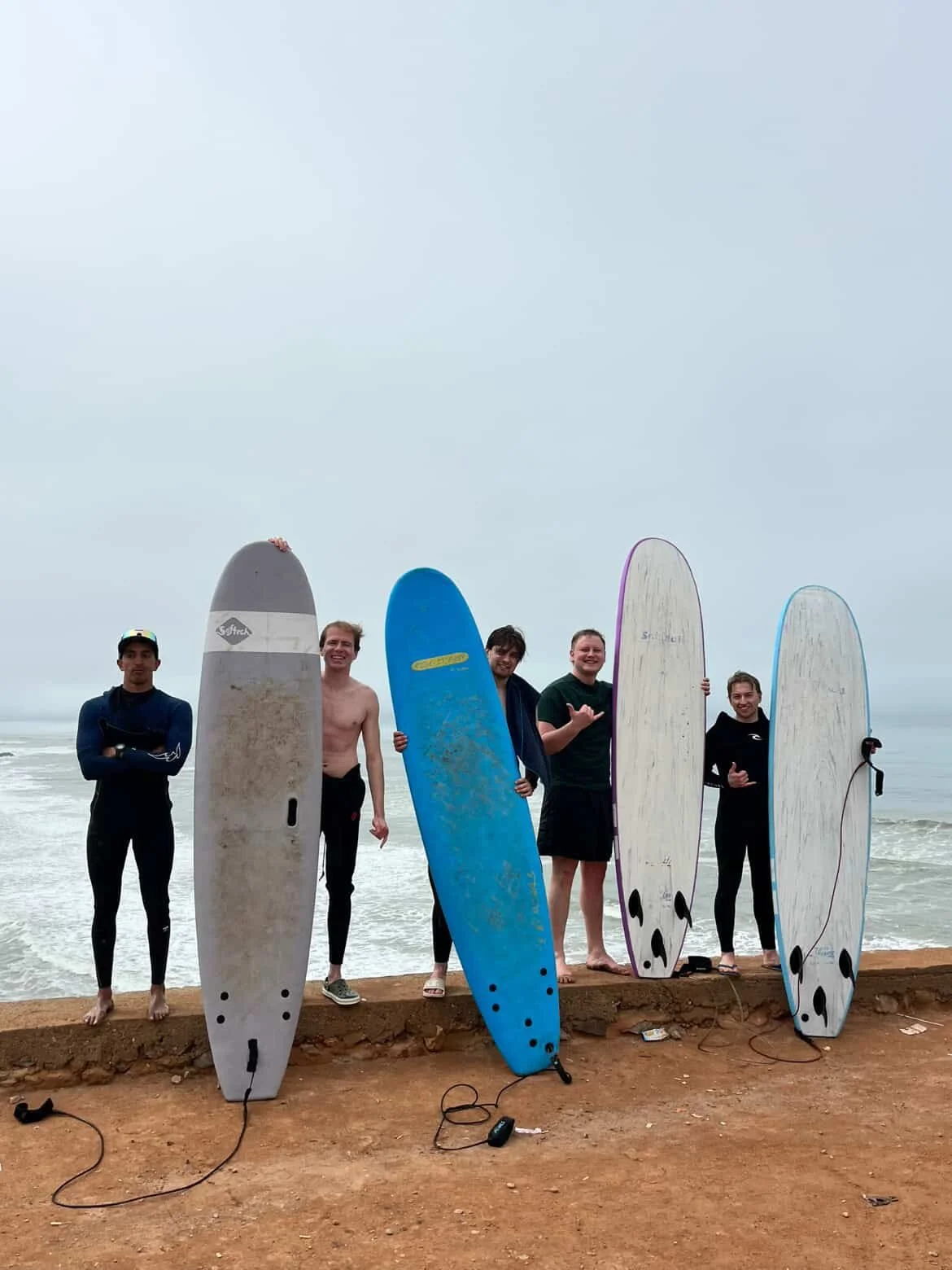 Group of five young men with surfboards on a beach during cloudy weather, some smiling and giving thumbs up.