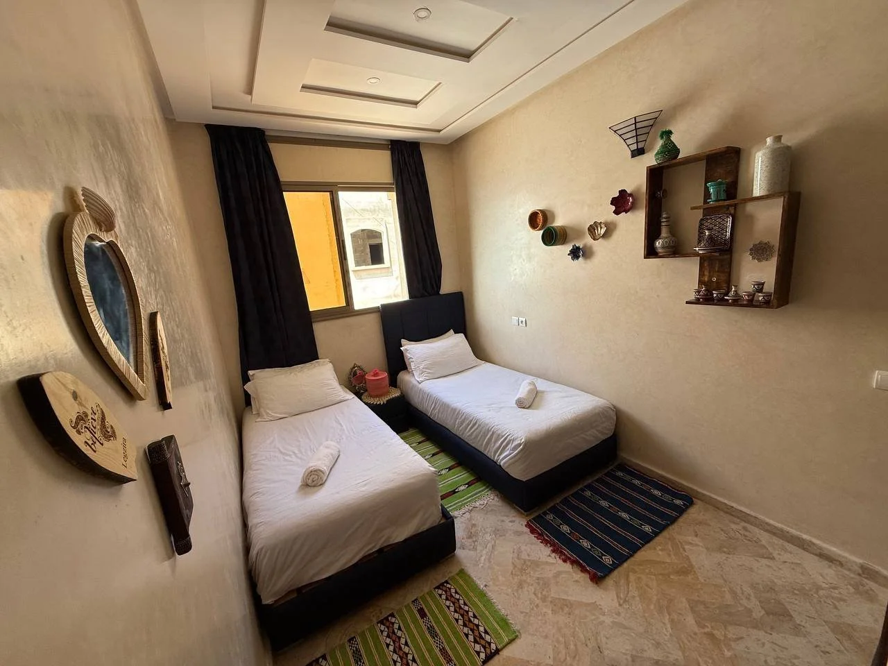 A bedroom with two single beds with white linens, rolled towels, and dark blue headboards. There are colored rugs on a marble floor, a window with black curtains, and decorative shelves with pottery on the beige wall.
