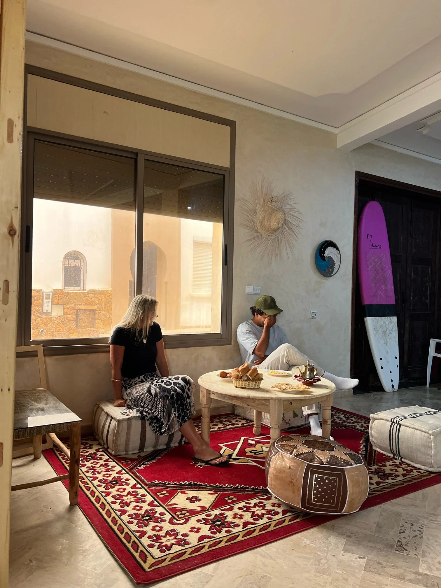 Two women sitting on cushions on a red patterned rug in a cozy living room. One woman is sitting on a low cushioned stool, the other on a floor cushion, with a wooden coffee table between them holding a tea set, bread, and plates. A surfboard is lean