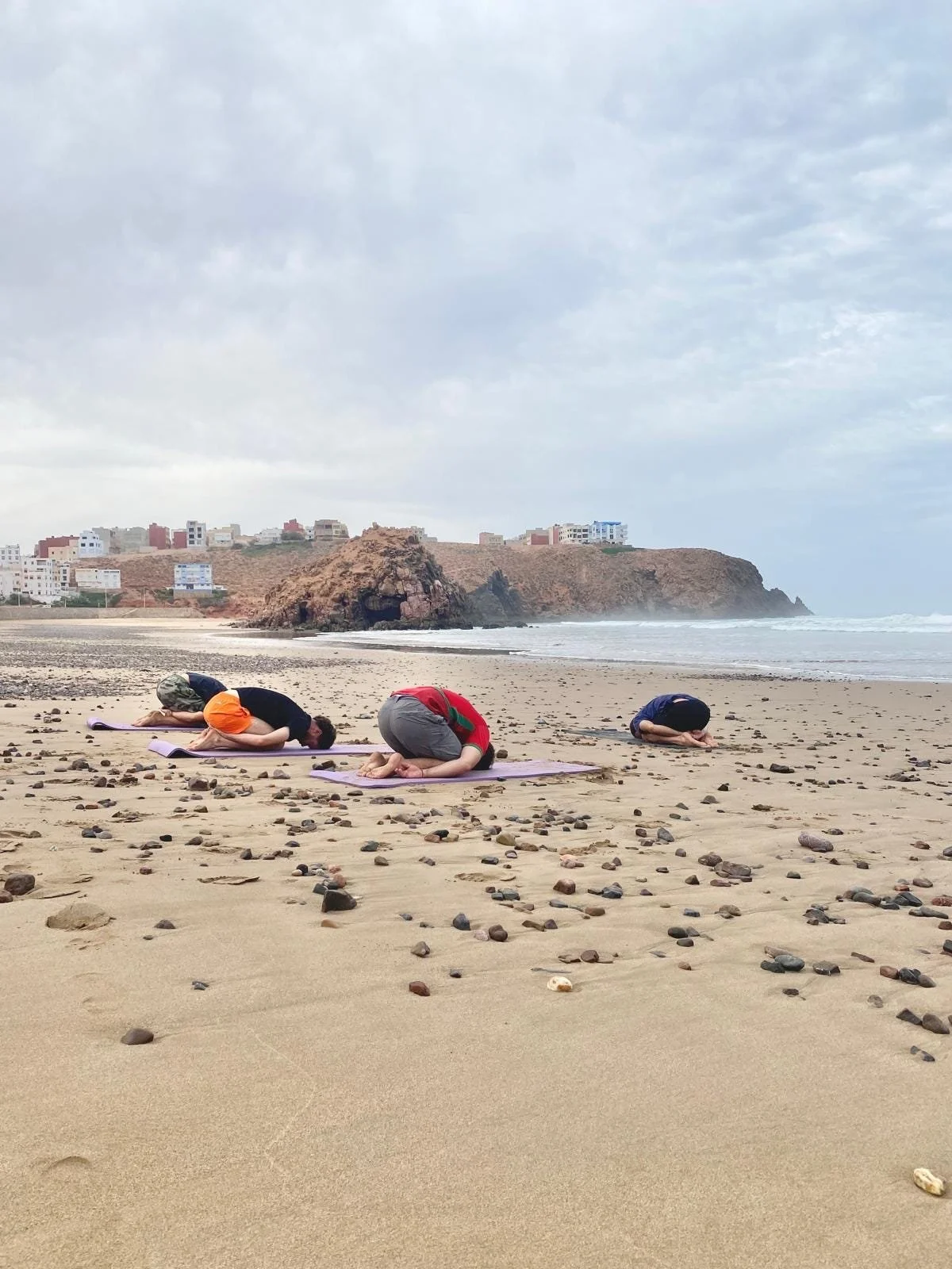Four people practicing yoga on the beach, in child's pose, with rocks scattered on sand, overlooking ocean and cliffs with buildings in background.