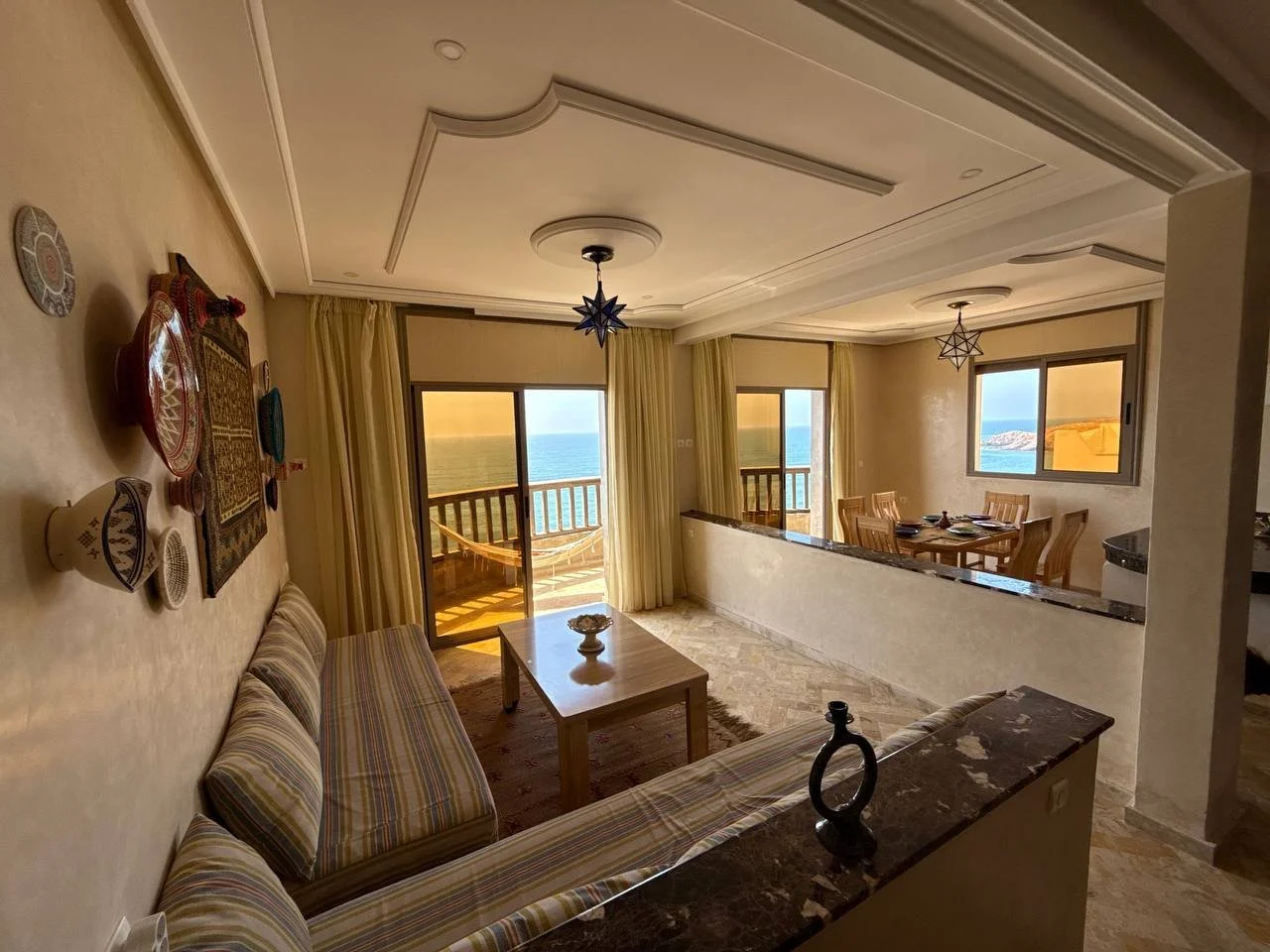 Living room with sliding glass doors opening to a balcony overlooking the ocean, decorated with patterned sofa, wooden dining table, and ocean view windows, soft lighting, ceiling beams, and hanging star-shaped lights.