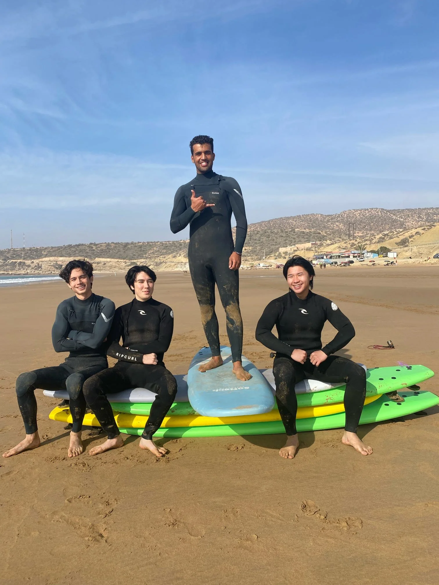 A group of five people at the beach with surfboards, one person standing on a surfboard, making a shaka sign, and four others sitting on surfboards, all wearing black wetsuits, with a hilly landscape and a partly cloudy sky in the background.