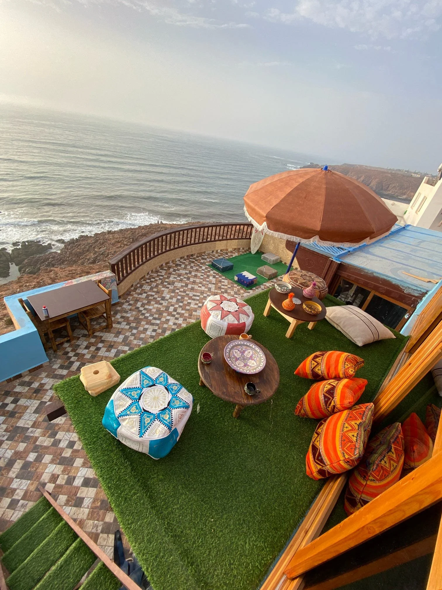 A terrace overlooking the ocean with a brown parasol, and colorful cushions on the floor, decorated with vibrant pillows, small tables, and a rug, with rocky coastline and the sea in the background.