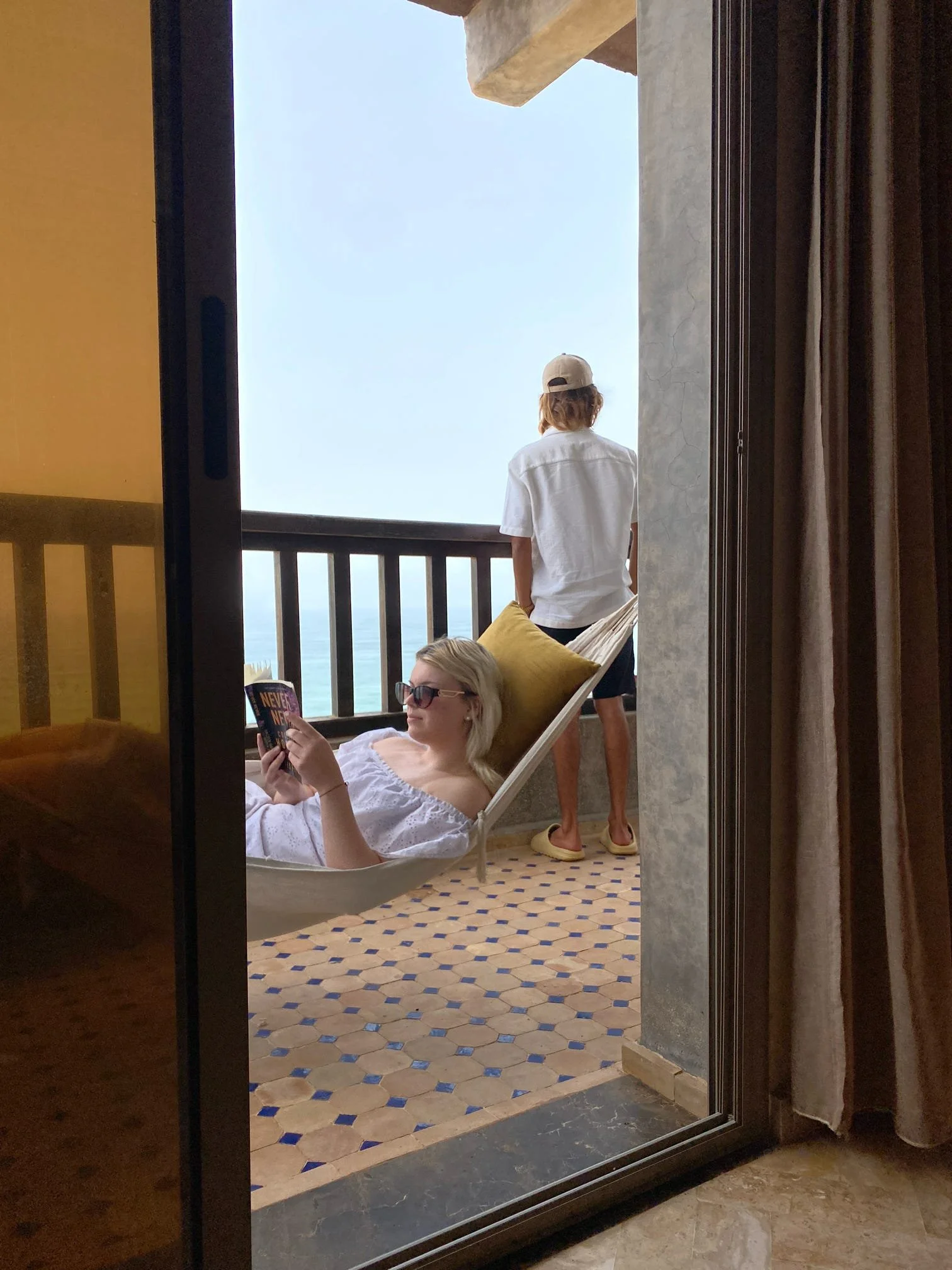 Woman with blonde hair, sunglasses, and white clothing relaxing on a hammock while reading a book. Behind her, a person with short hair, wearing a white shirt, beige cap, and yellow slippers, stands on a balcony overlooking a body of water.