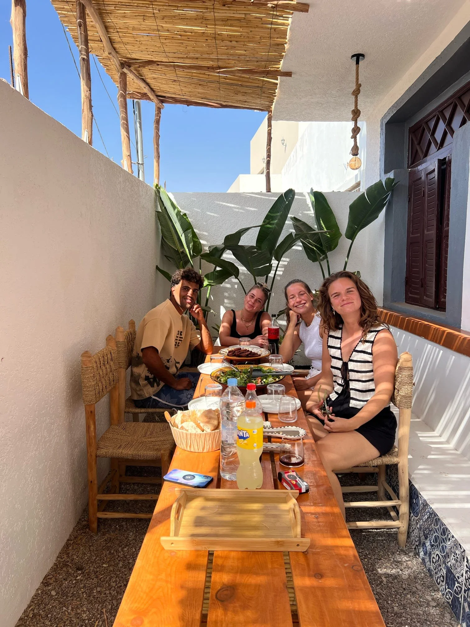 Five young adults sitting at a long wooden table outdoors, enjoying a meal. The setting features white walls, large green tropical plants, and a sunny sky. On the table, there are various dishes, drinks, and snacks.